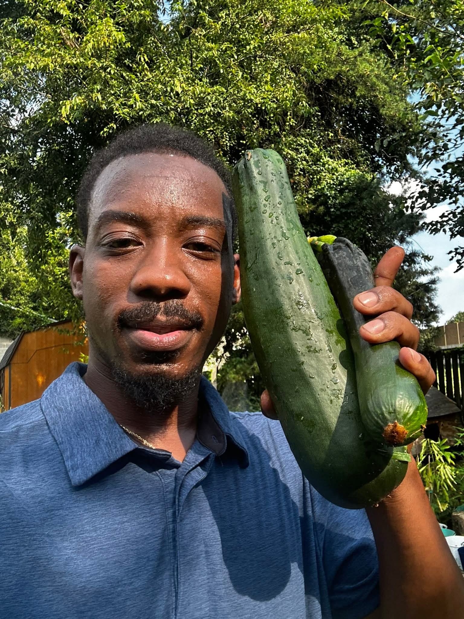 Young man holding two large zucchinis outdoors with trees in background, smiling, wearing a blue shirt.