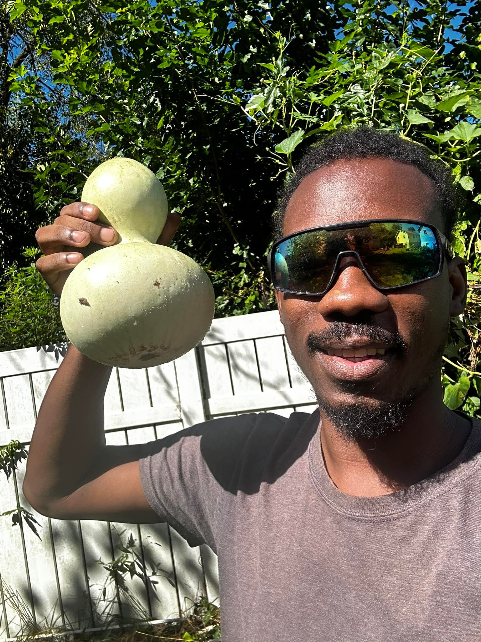 Man with sunglasses holding a large gourd outdoors, surrounded by green foliage, smiling.