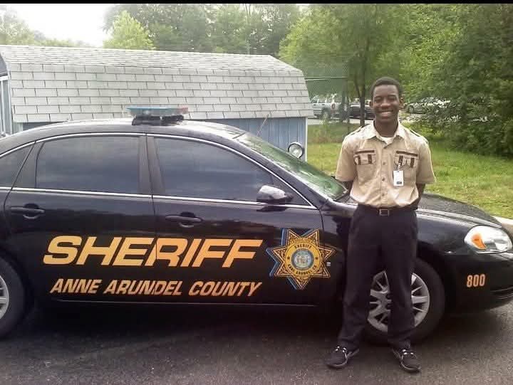 Young man in sheriff uniform standing next to sheriff vehicle outdoors, trees and building in background.
