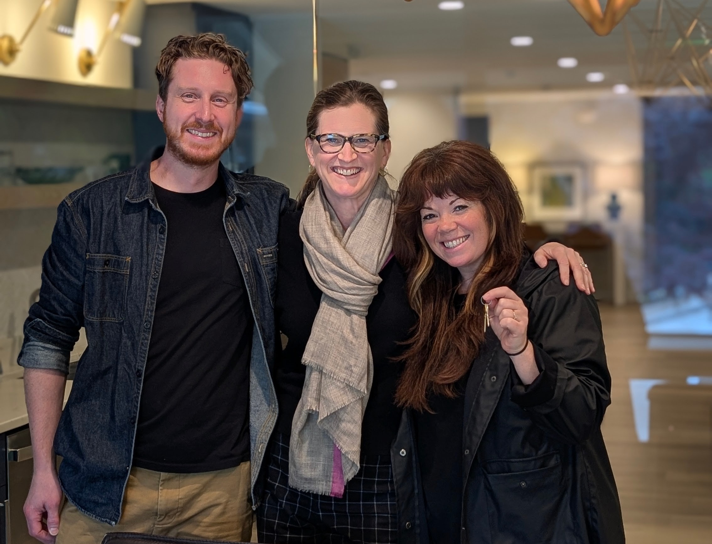 Three people smiling indoors, two women and one man, standing close together with arms around each other.