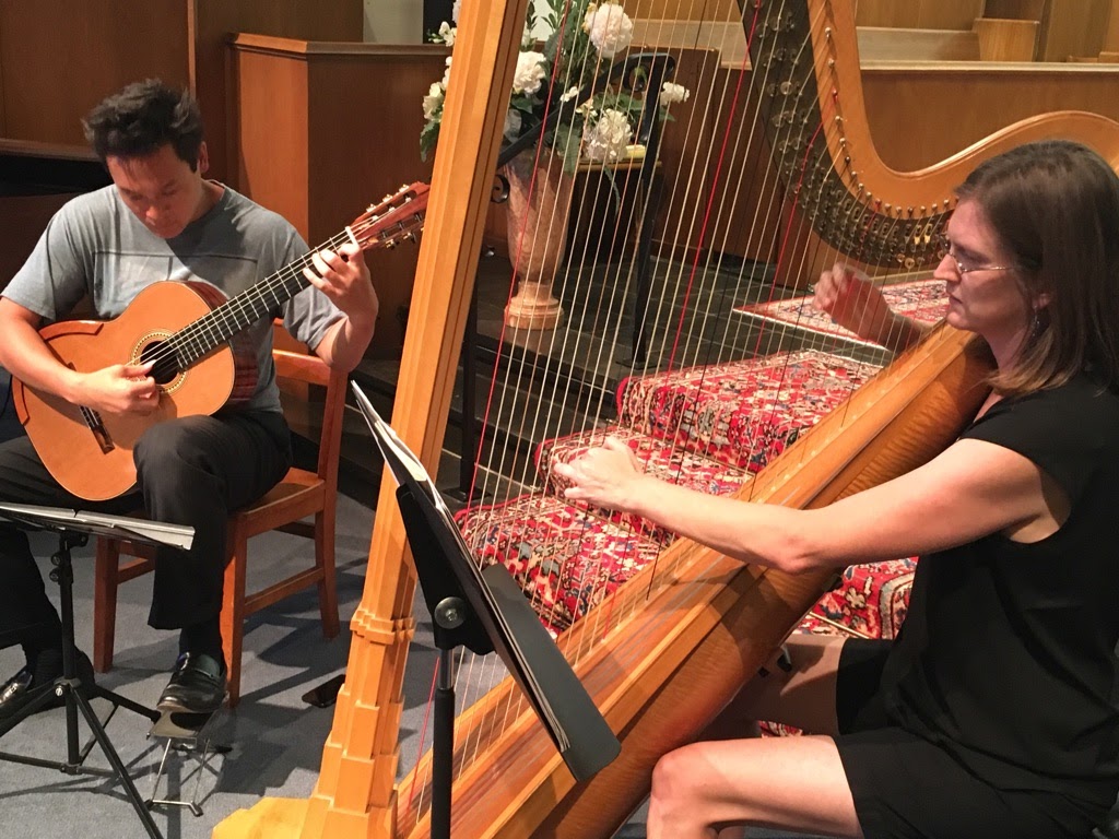 Two women playing musical instruments: one with a guitar, the other with a harp, in a room with wooden furniture.