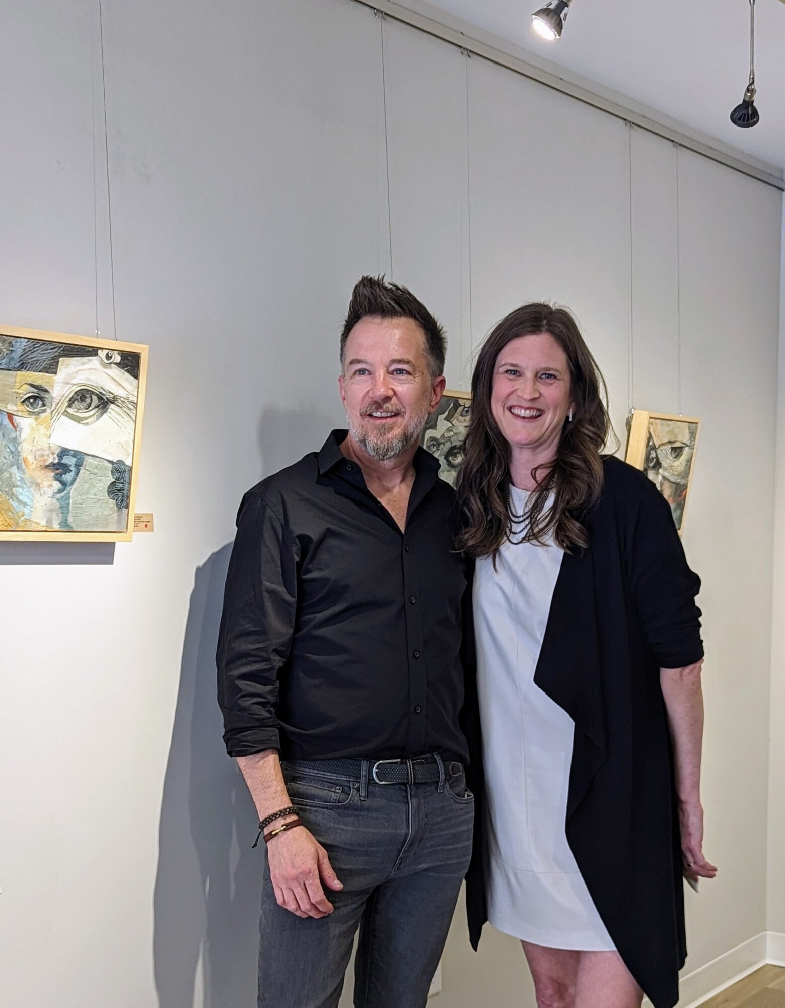 Two people, a man and a woman, stand together smiling in an art gallery with paintings on the wall.