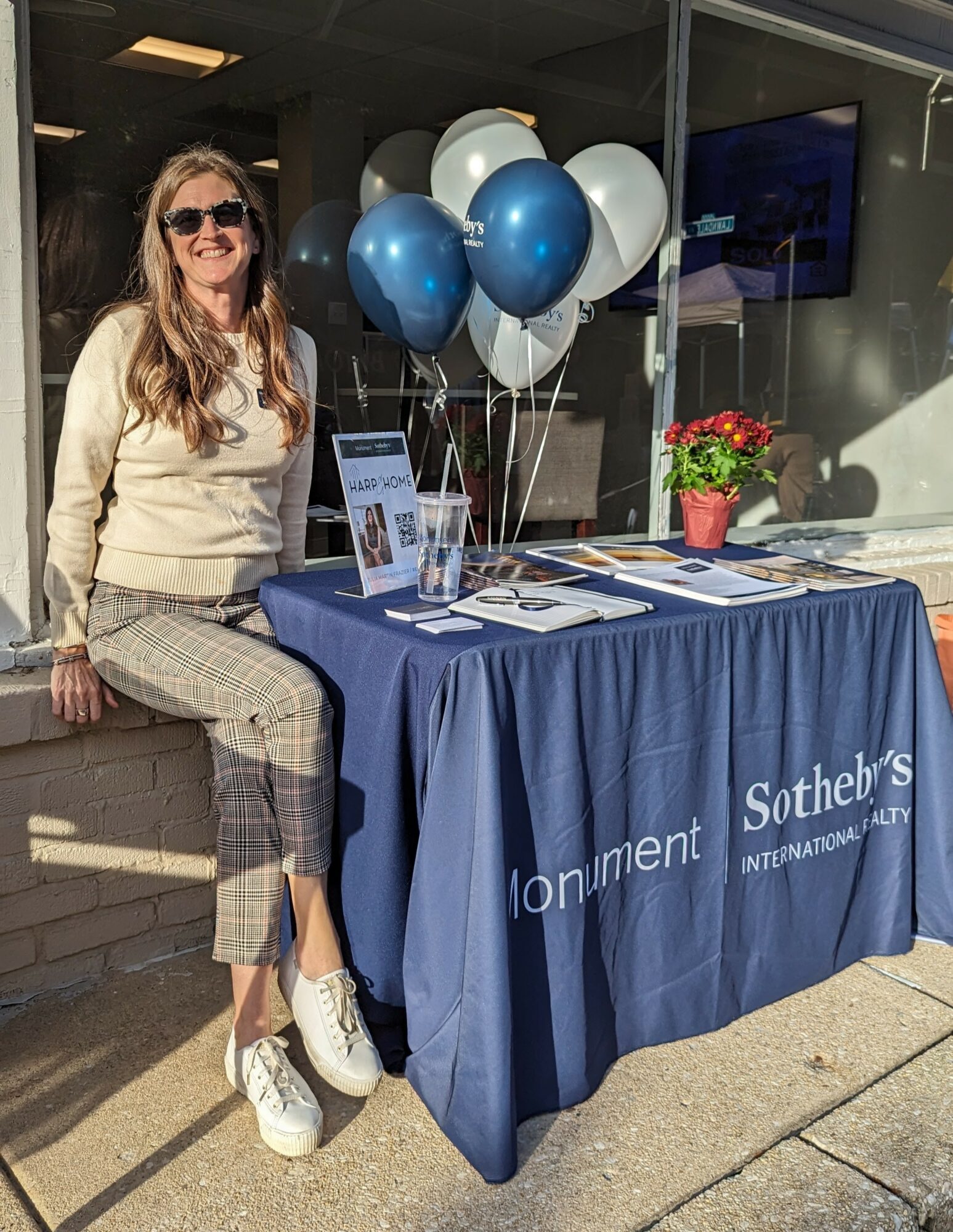 Woman sitting at a table with balloons and brochures outside, sunlight casting shadows, table covered with a blue cloth, potted plant nearby.