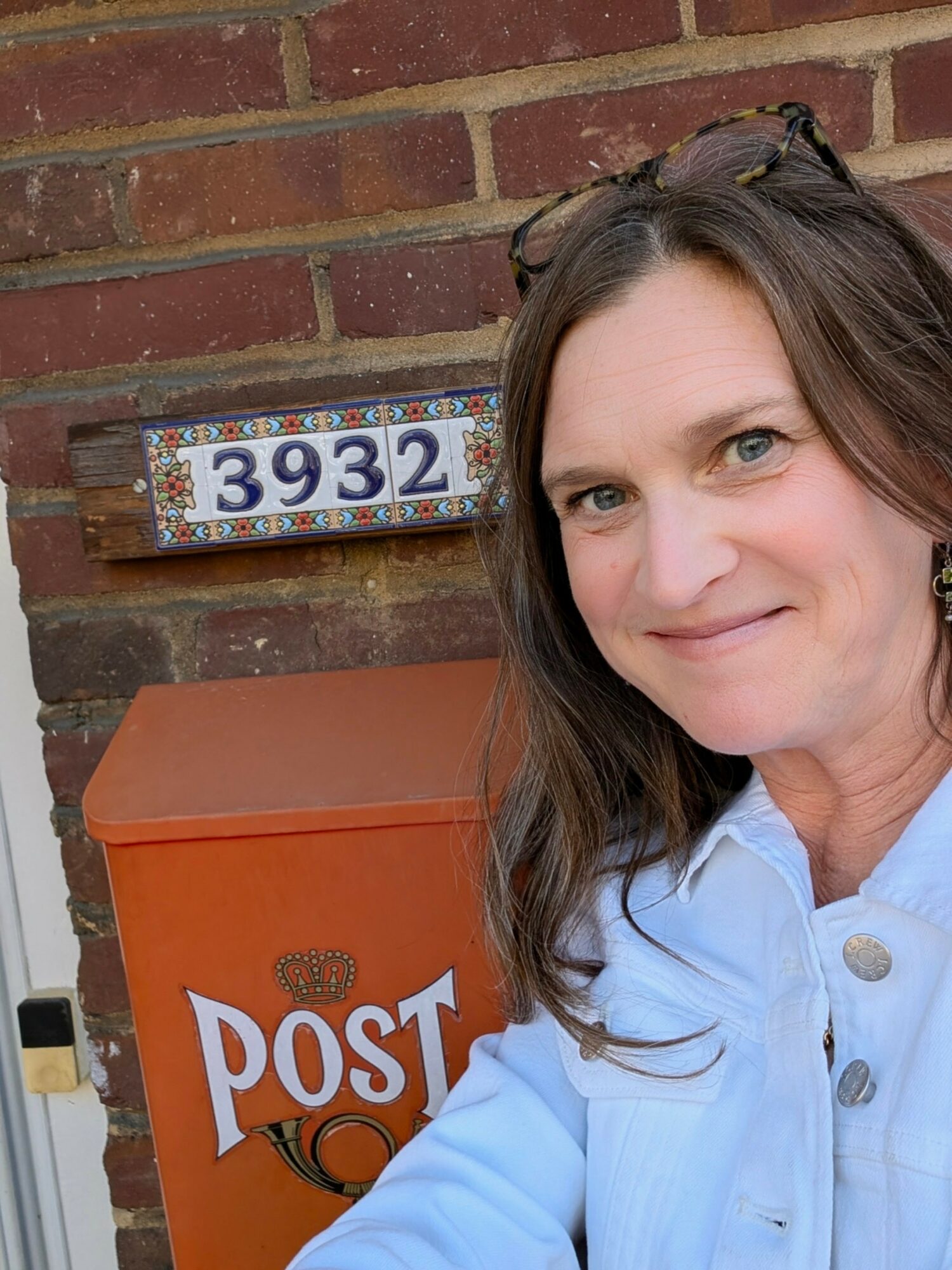 Woman with brown hair and glasses on head, smiling, standing next to a brick wall and orange mailbox with 'POST' written on it.