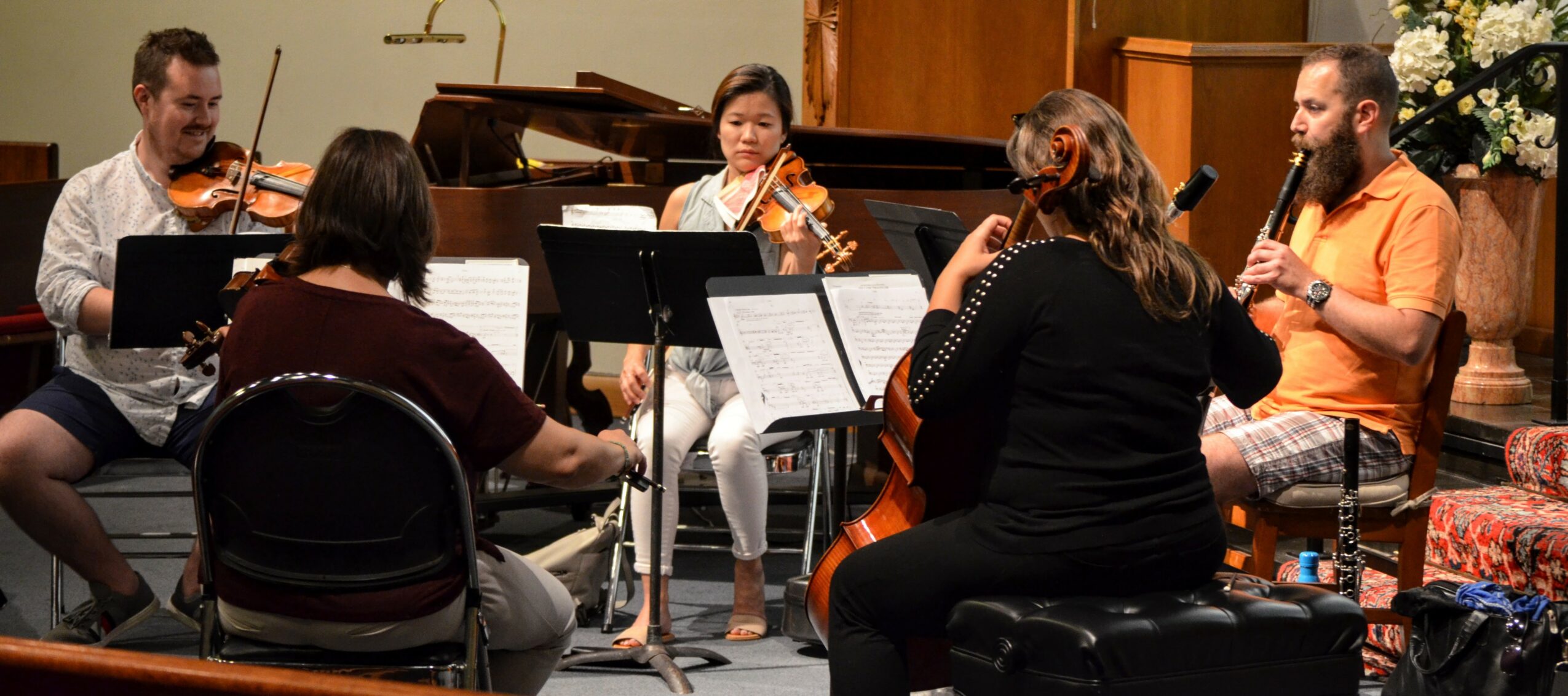 Group of six people playing musical instruments in a room, with music stands and a piano, engaged in a rehearsal or performance.
