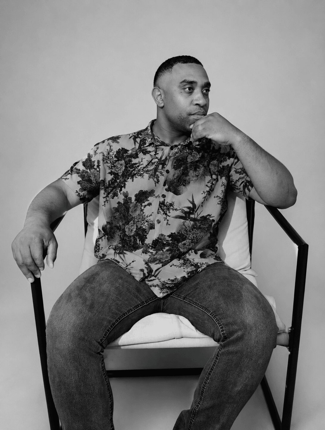 Man sitting on a chair with one hand on his chin, wearing a patterned shirt and jeans, against a plain background.