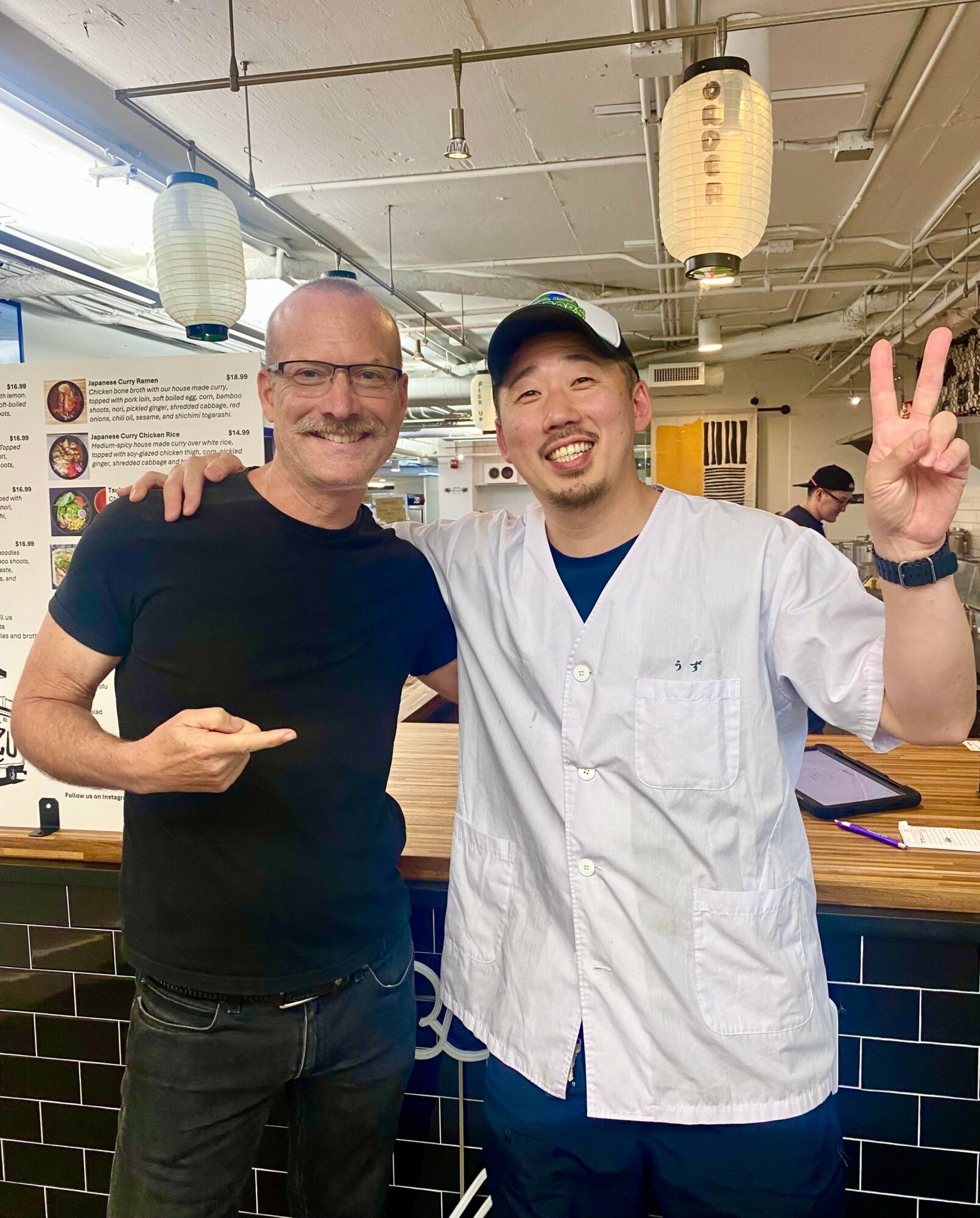Two men smiling, one in a black shirt and glasses, the other in a white chef's coat and cap, making a peace sign.