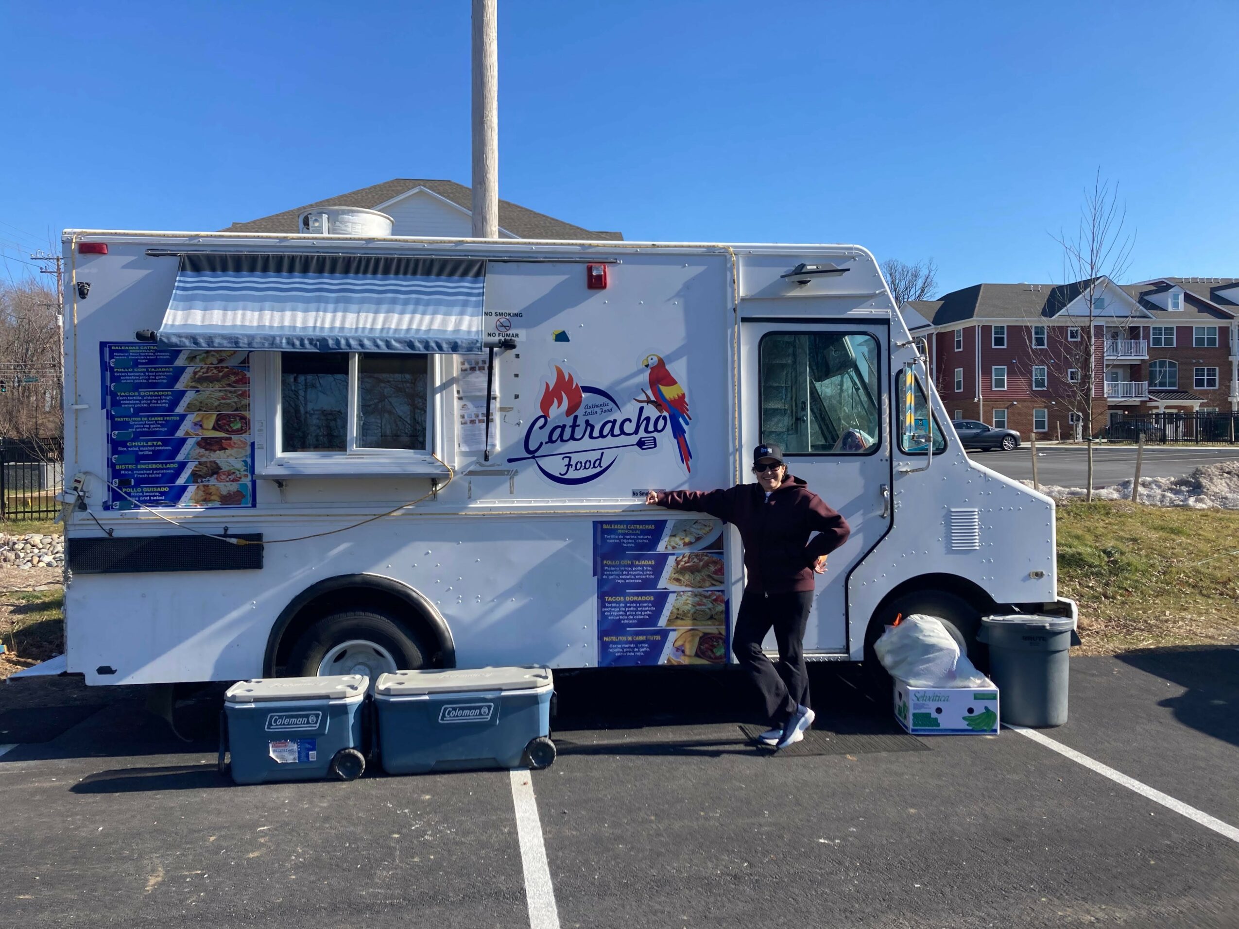 Person standing next to a food truck with a blue sky background, parked on a street with buildings in the distance.