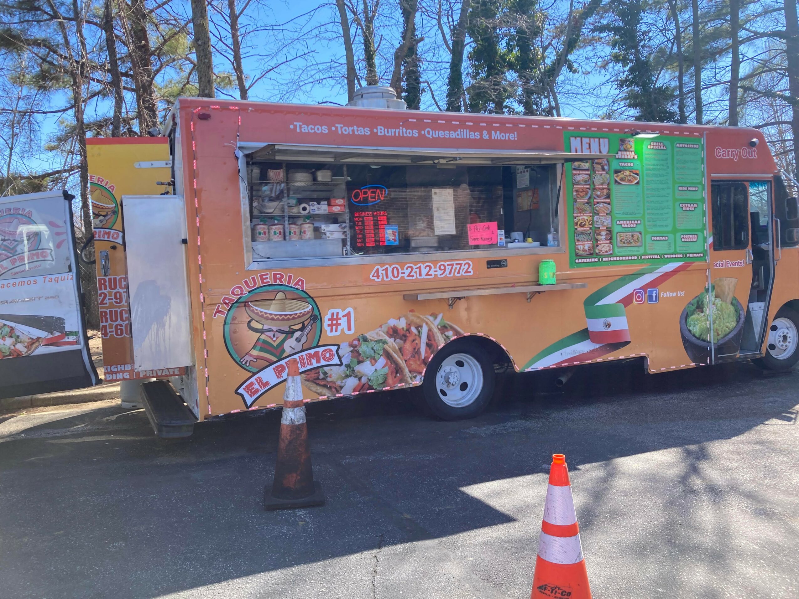 Food truck parked outdoors with trees in background, orange and green colors, serving window, and traffic cones nearby.