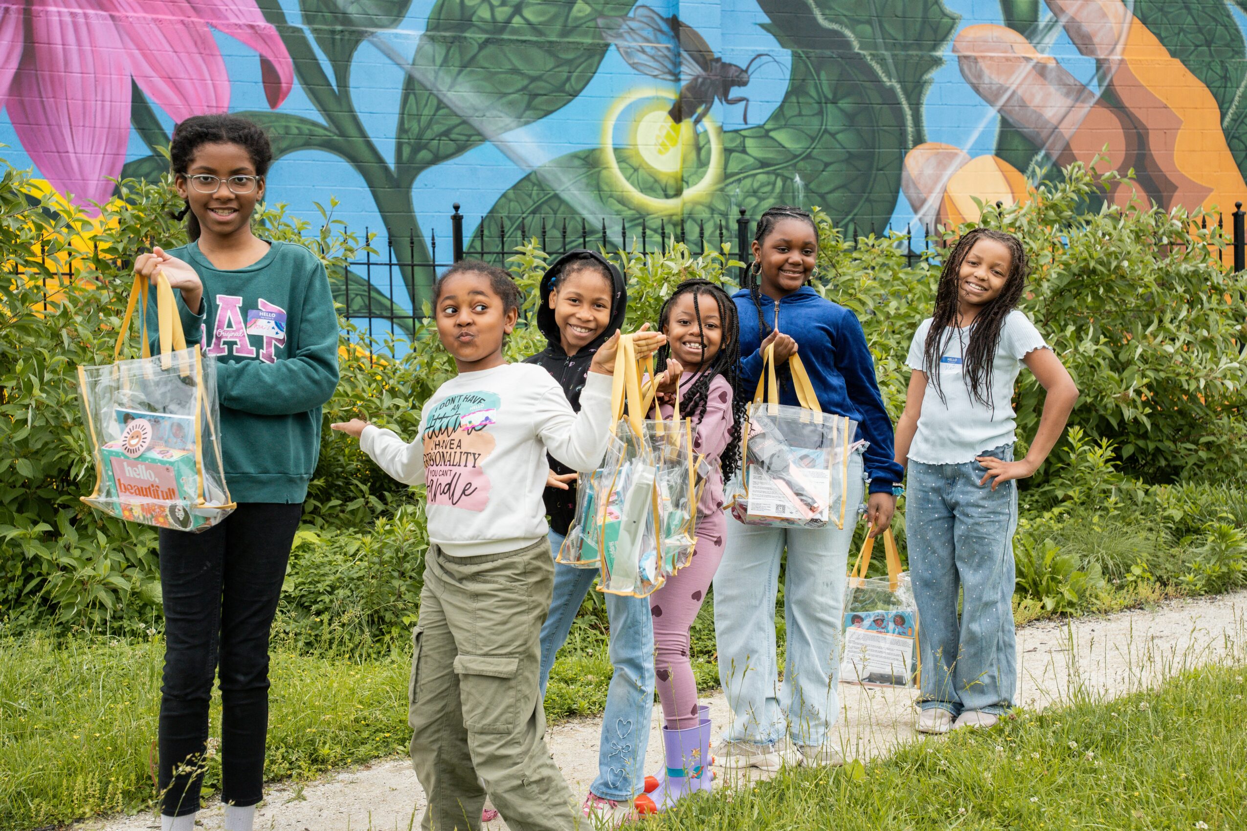 Six children standing outdoors in front of a colorful mural, holding bags and smiling.