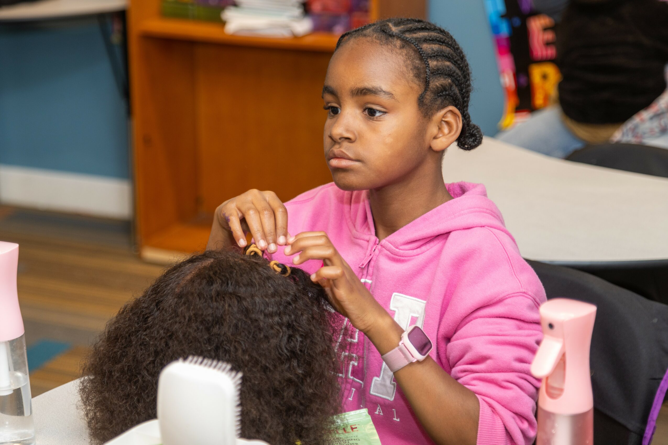 Young girl in pink hoodie sitting at a table with a person with curly hair, in a classroom setting.