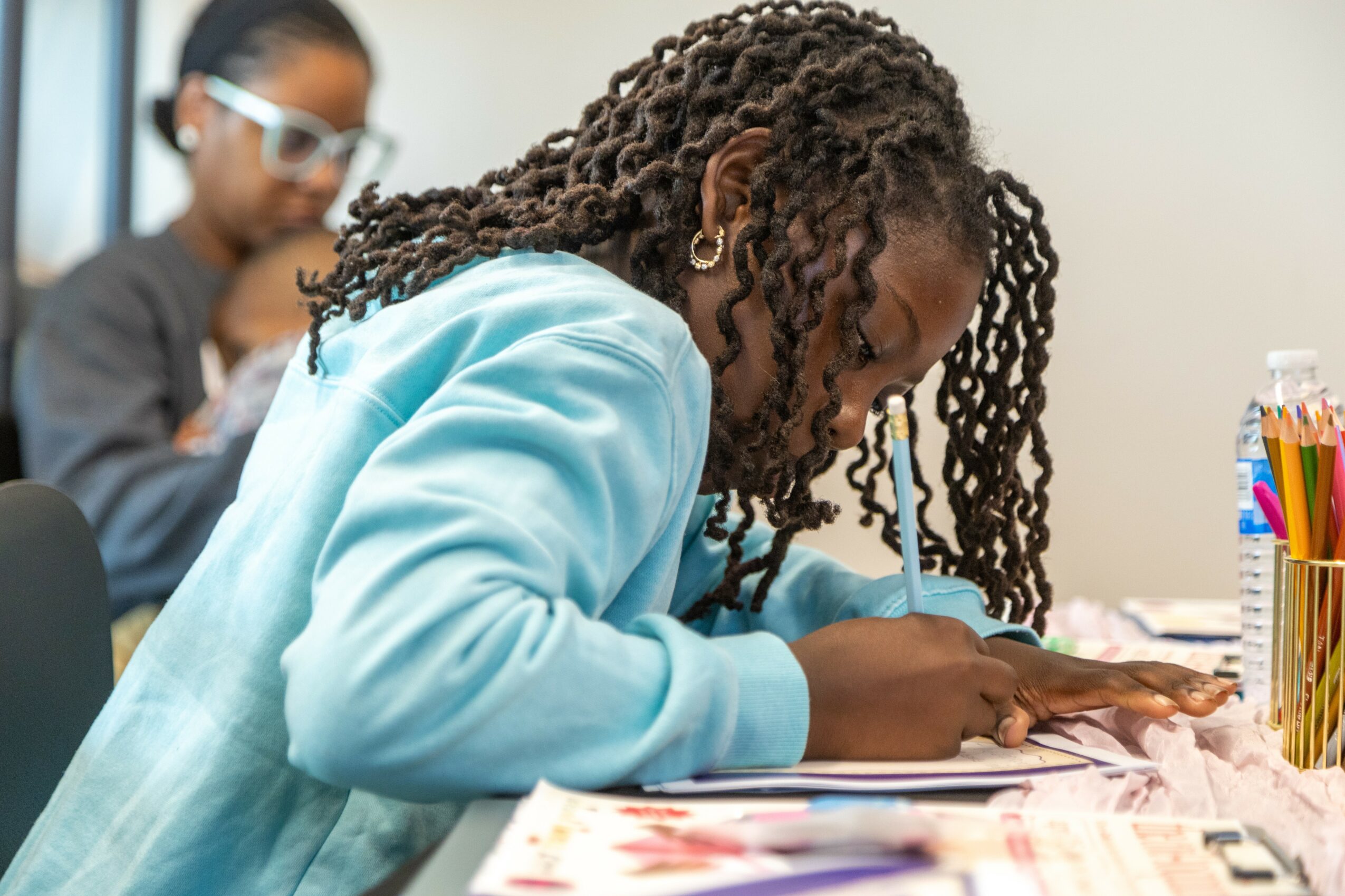 Girl with curly hair writing on paper at a desk, with another girl in the background, in a classroom setting.
