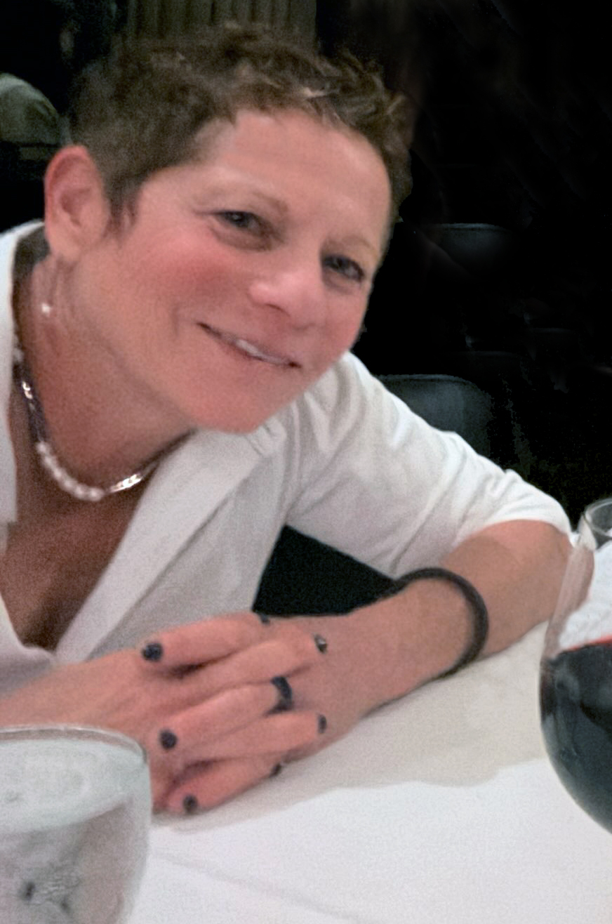 Woman with short hair smiling, wearing a white top and pearl necklace, sitting at a table.