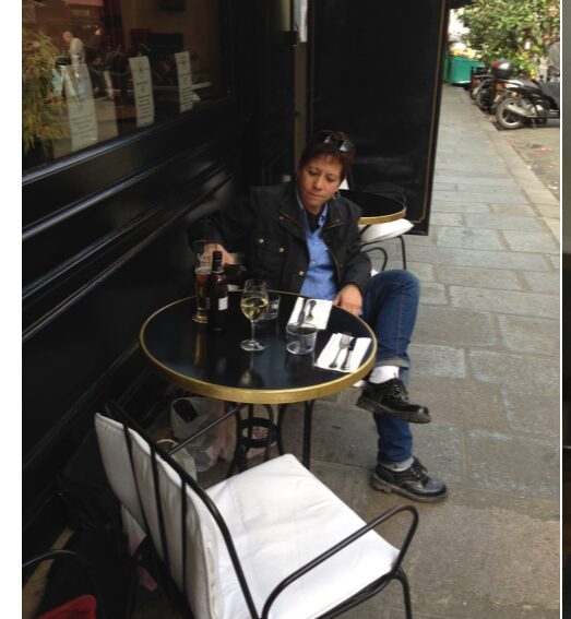 Person sitting at outdoor table with drinks, legs crossed, on a city sidewalk, wearing a denim jacket and jeans.