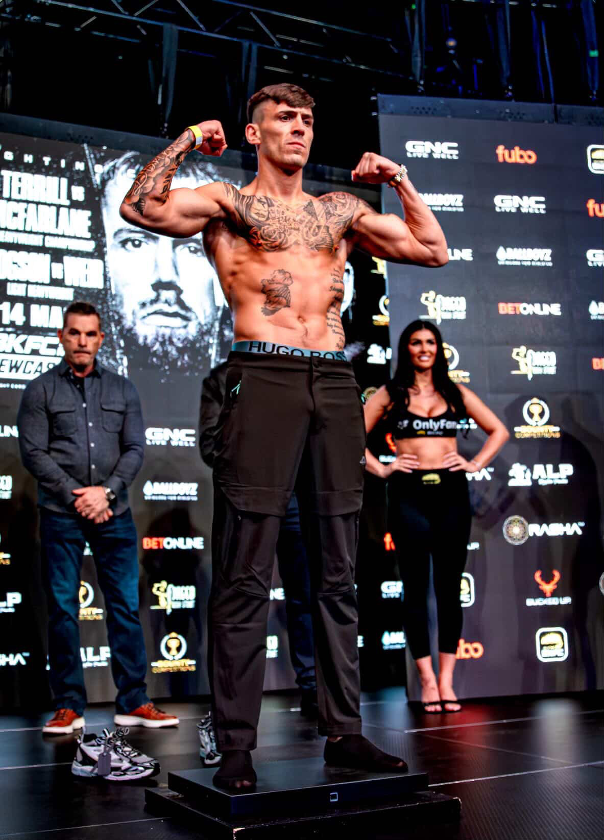 Male boxer with tattoos flexing arms on stage, two people behind him, event backdrop with logos and text, stage lighting.