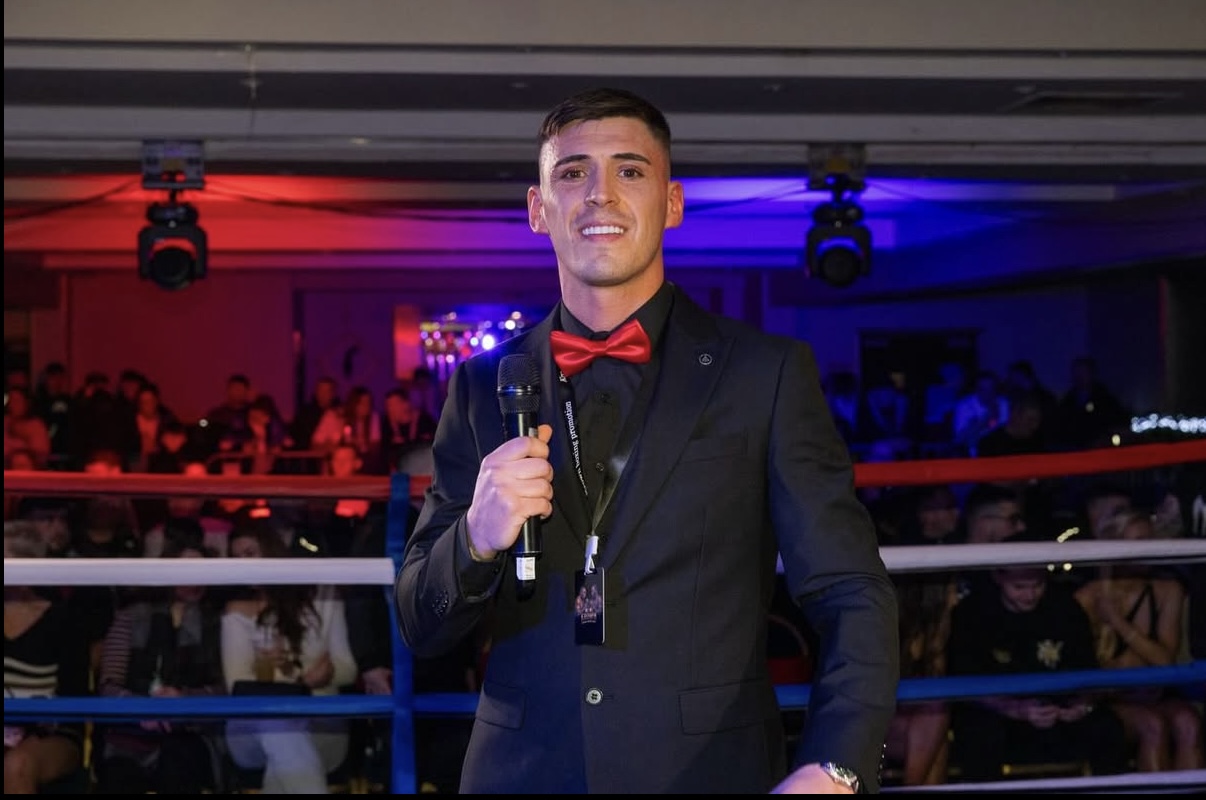 Man in tuxedo with red bow tie holding a microphone in a boxing ring, audience in background, colorful lighting