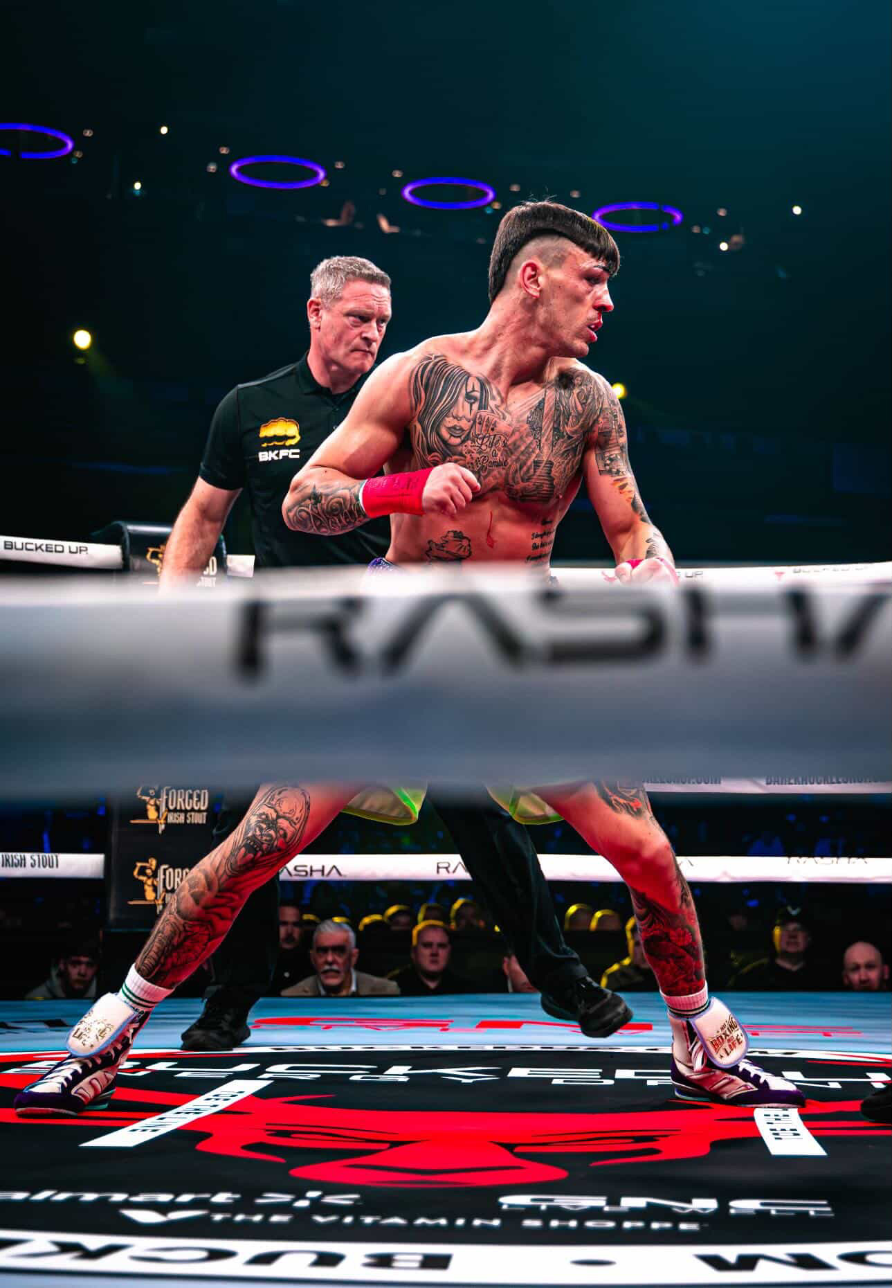 Boxer with tattoos and red gloves in ring, referee behind, audience in background, ring ropes visible.