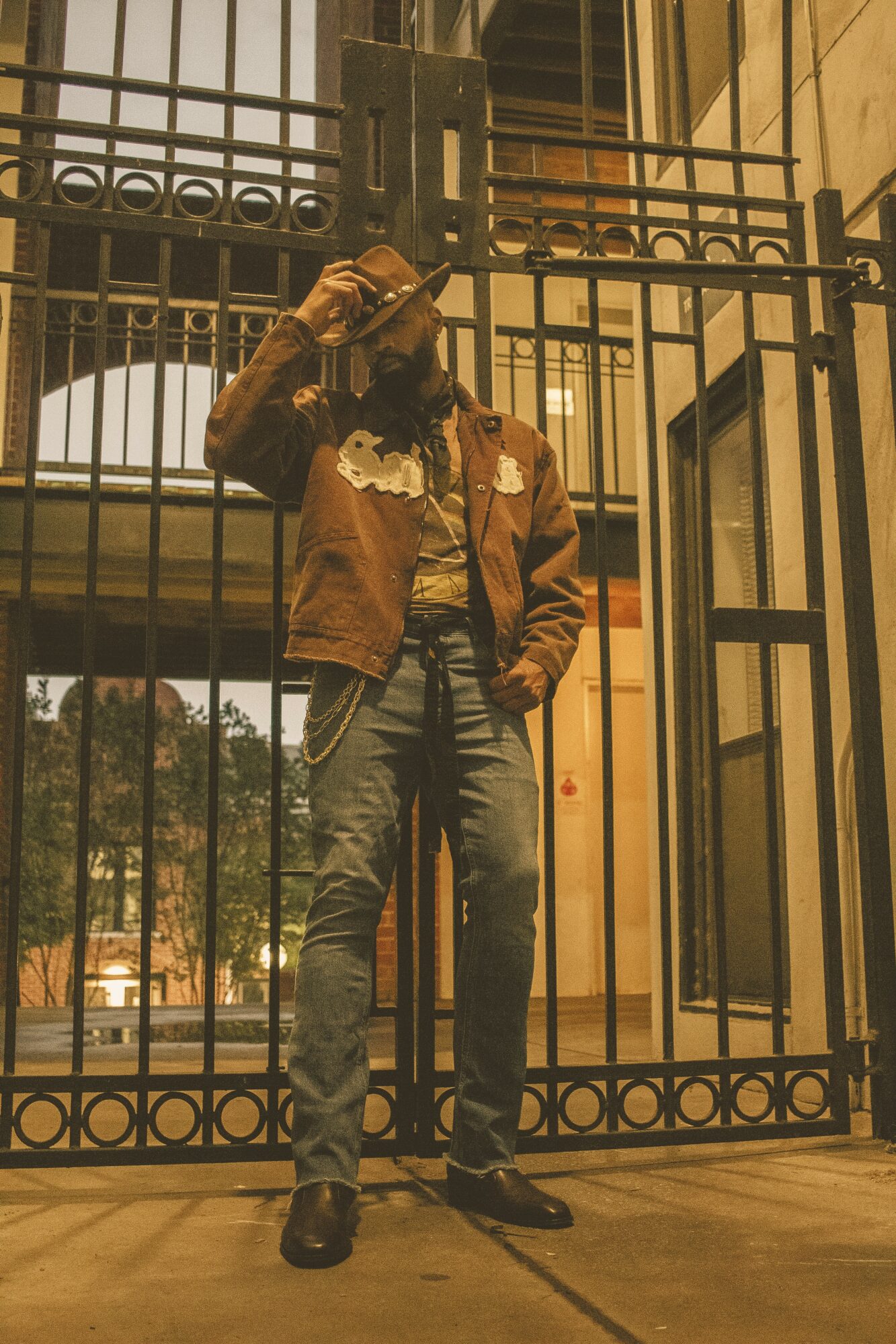 Person standing behind a metal gate, holding a phone to their ear, wearing a brown jacket and jeans.