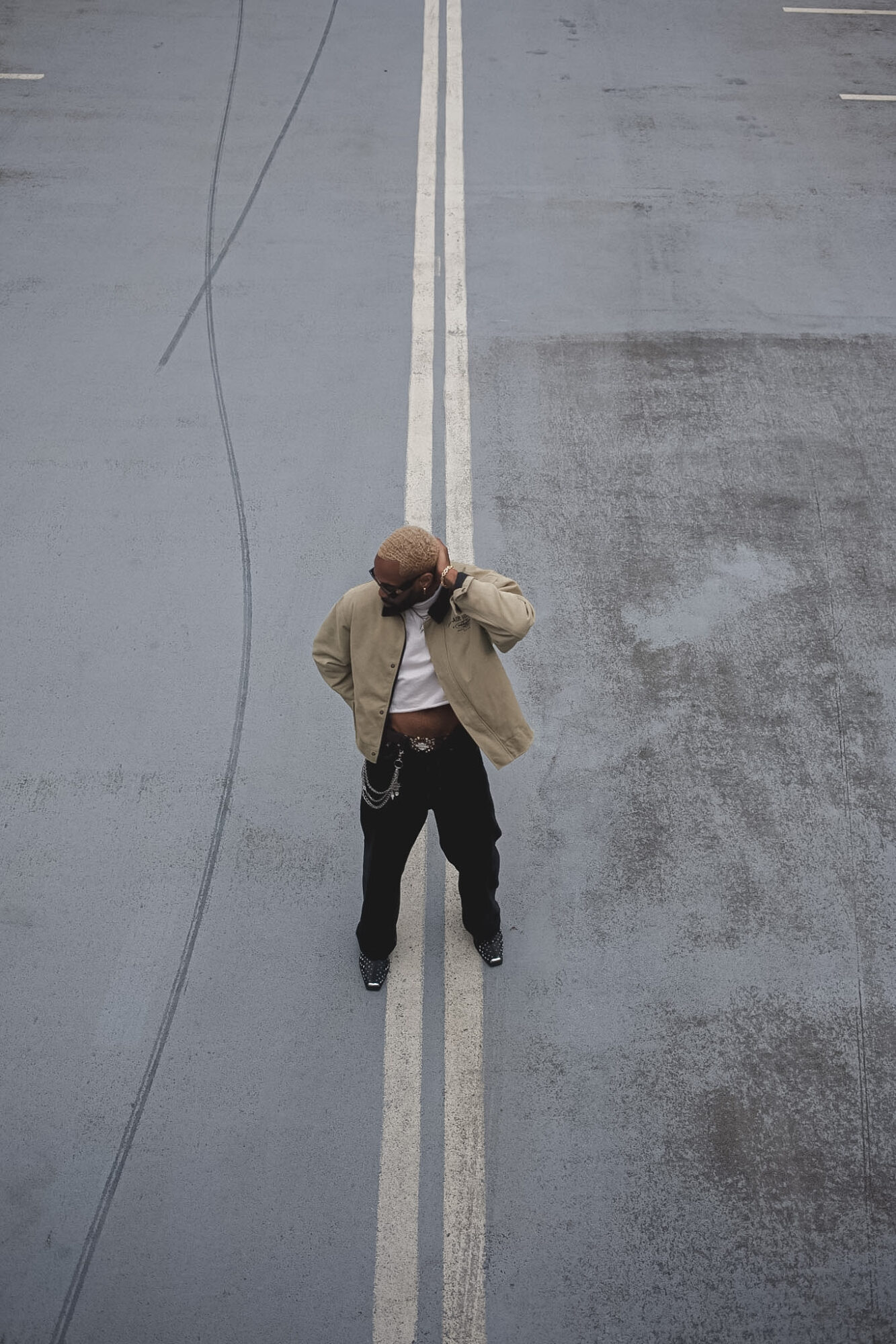 Person standing in the middle of a double yellow line on an empty road, looking down, with a beige jacket and dark pants.