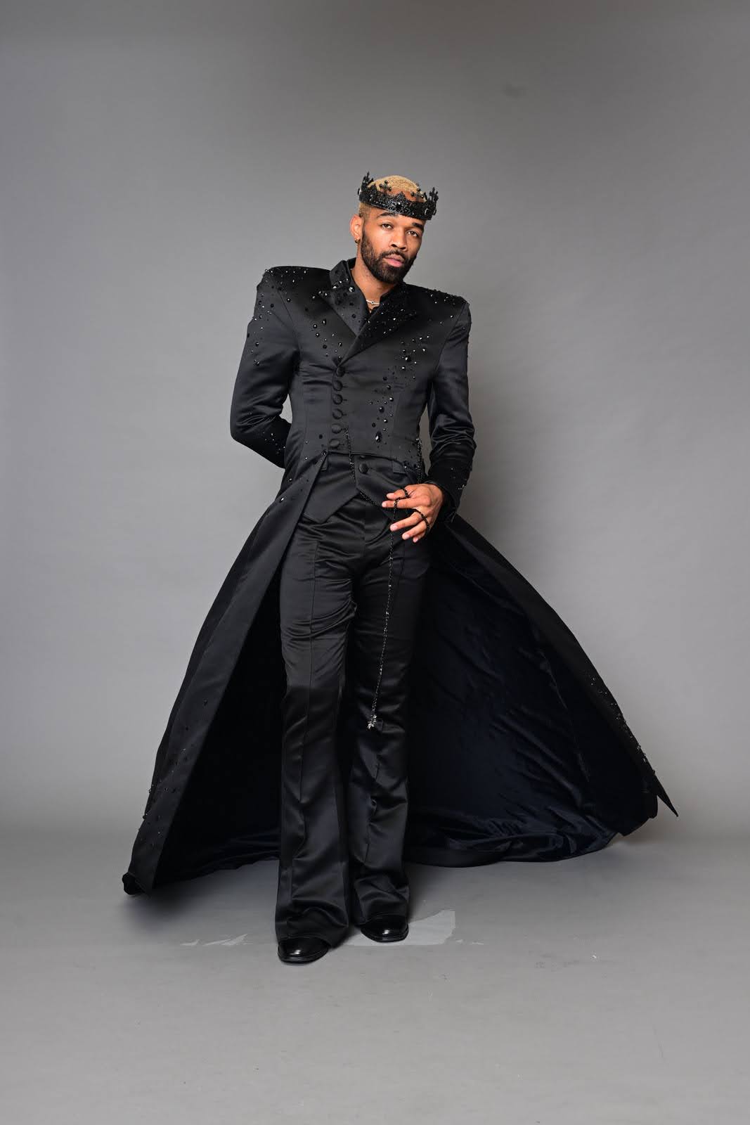 Man in black outfit with a long coat, standing against a plain background, looking at the camera.
