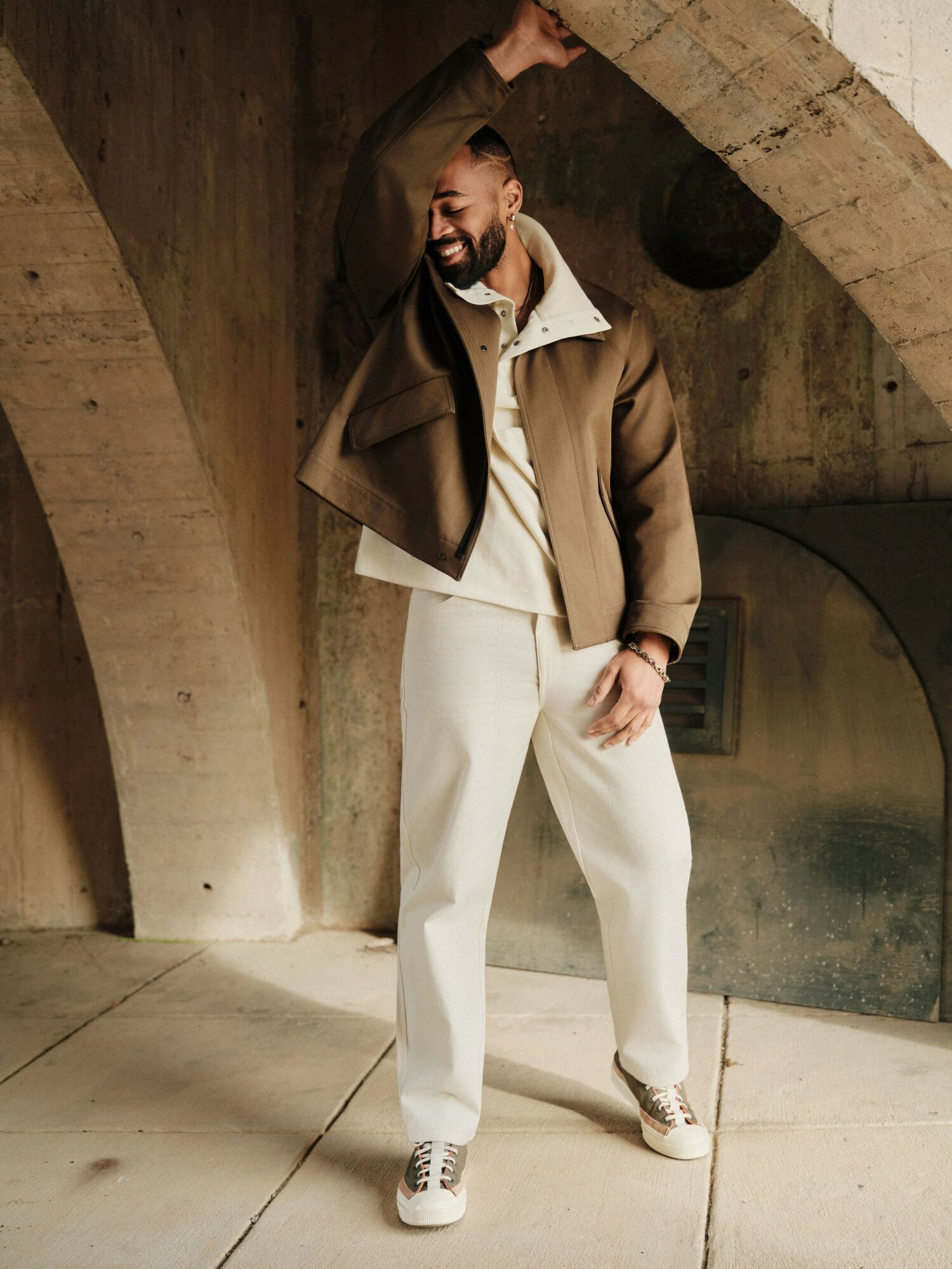 Man smiling, standing under concrete arch, wearing beige jacket, white pants, and sneakers, with one arm raised.