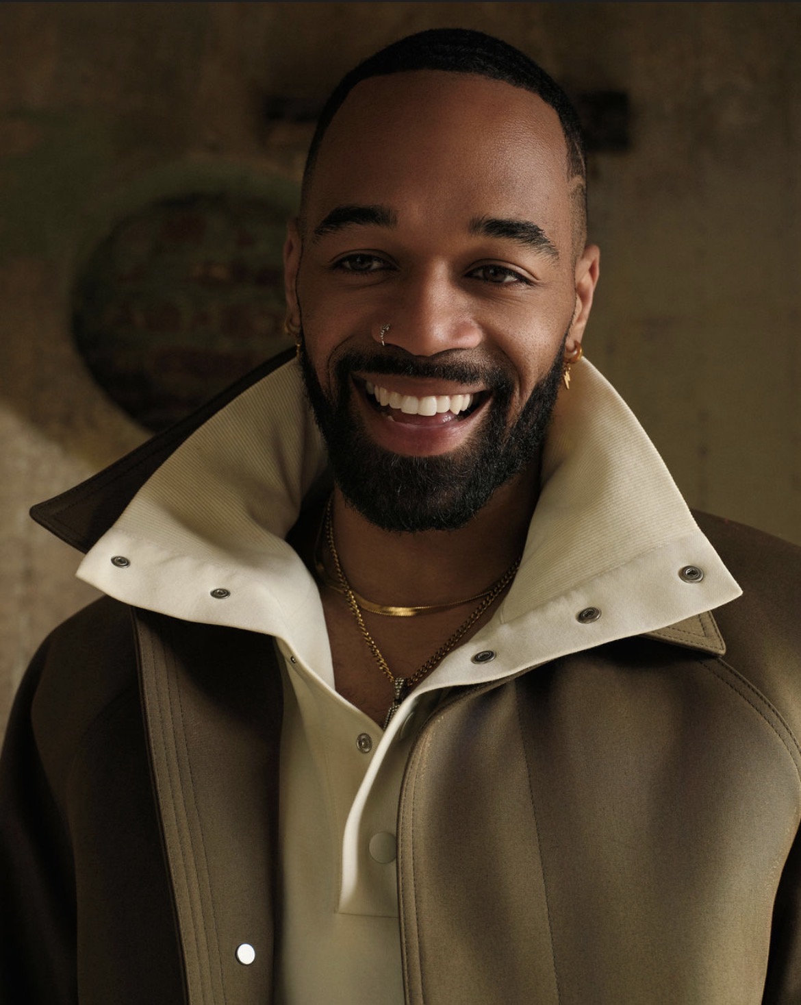Smiling man with a beard wearing a jacket and gold jewelry, looking to the side.