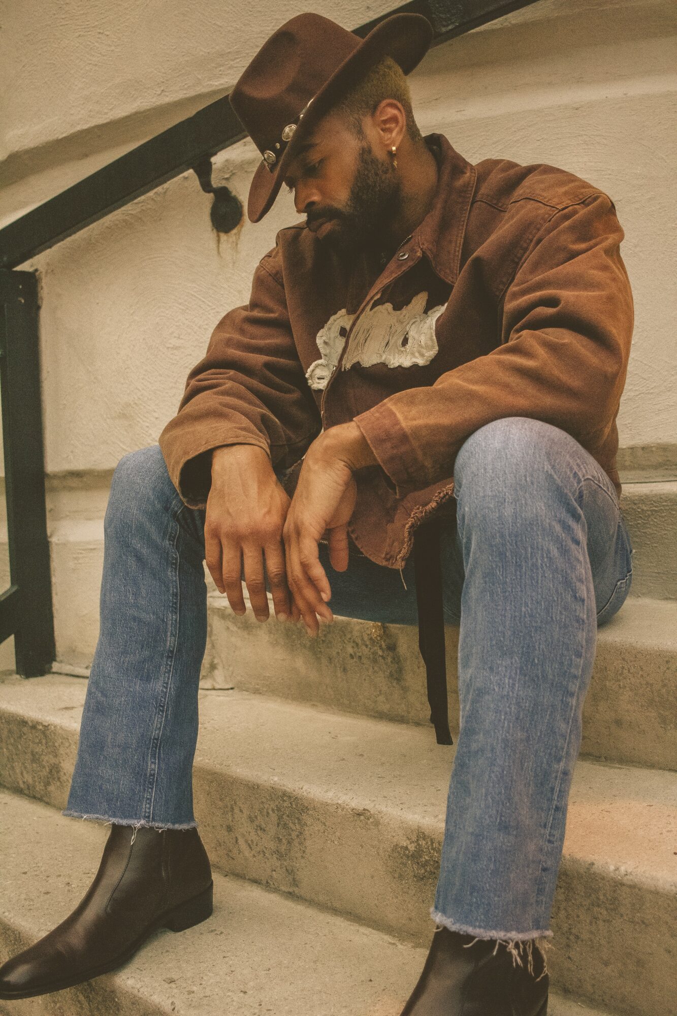 Man sitting on stairs wearing a cowboy hat, brown jacket, jeans, and black boots, looking down with hands clasped.