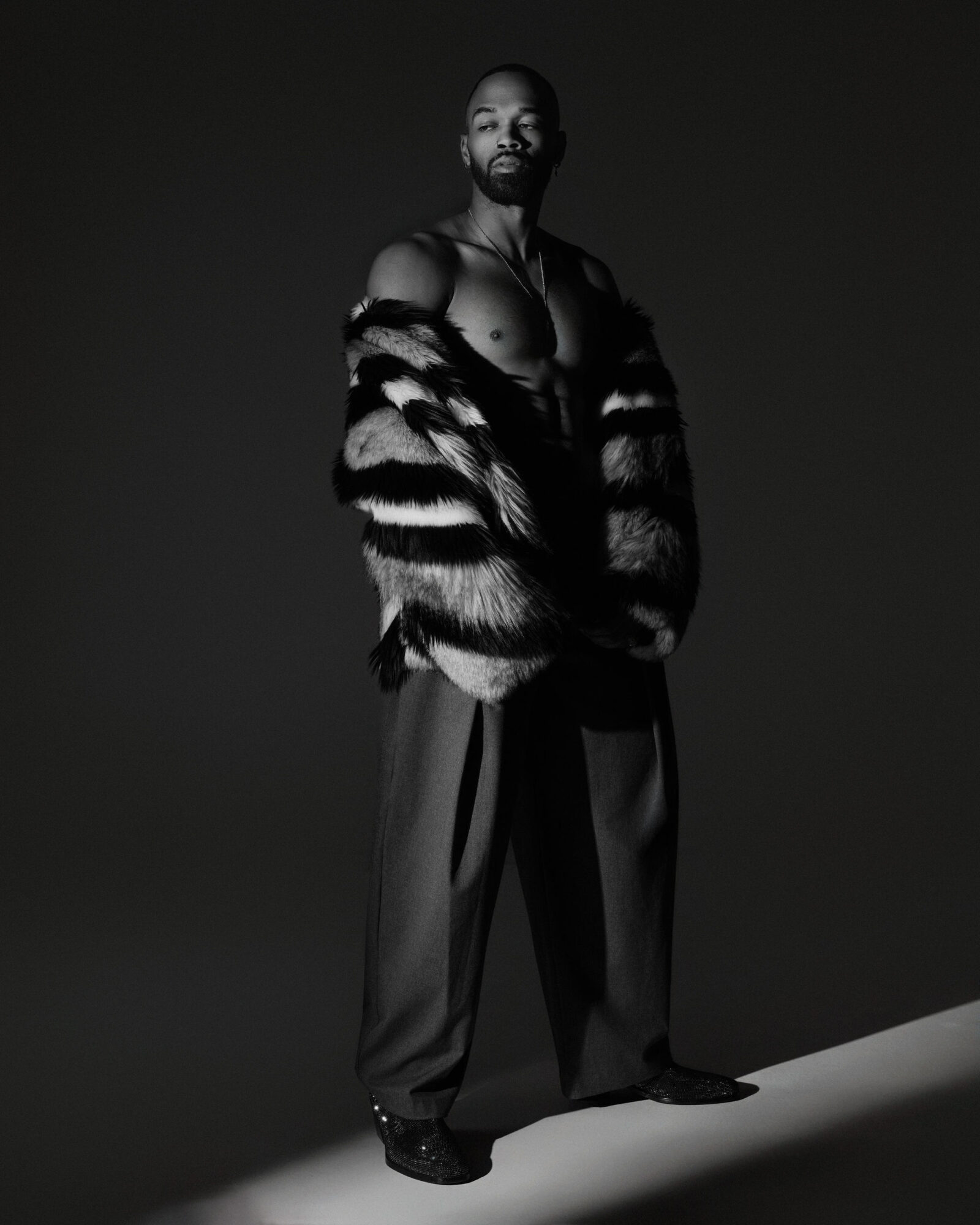 Man standing shirtless with a fur coat draped over shoulders, dark background, looking at camera, confident pose.