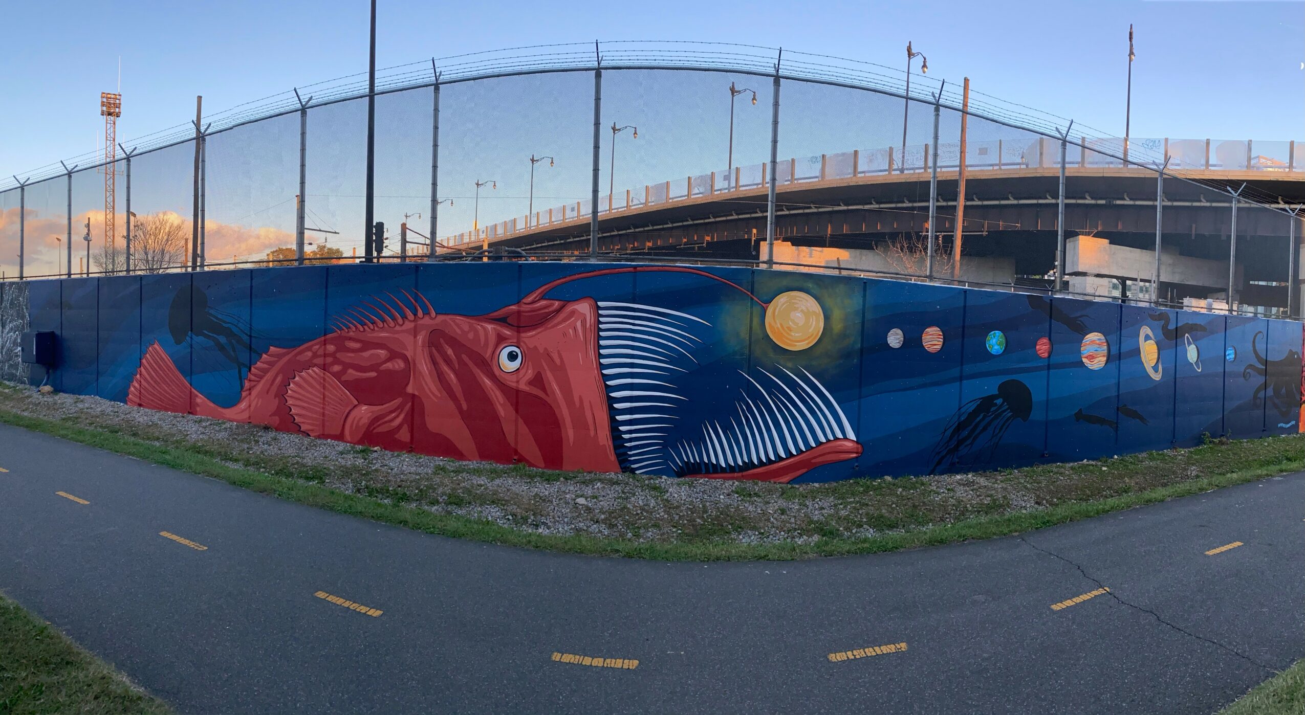 Colorful mural of a red fish with blue and white stripes on a blue background, painted on a wall near a sports field.
