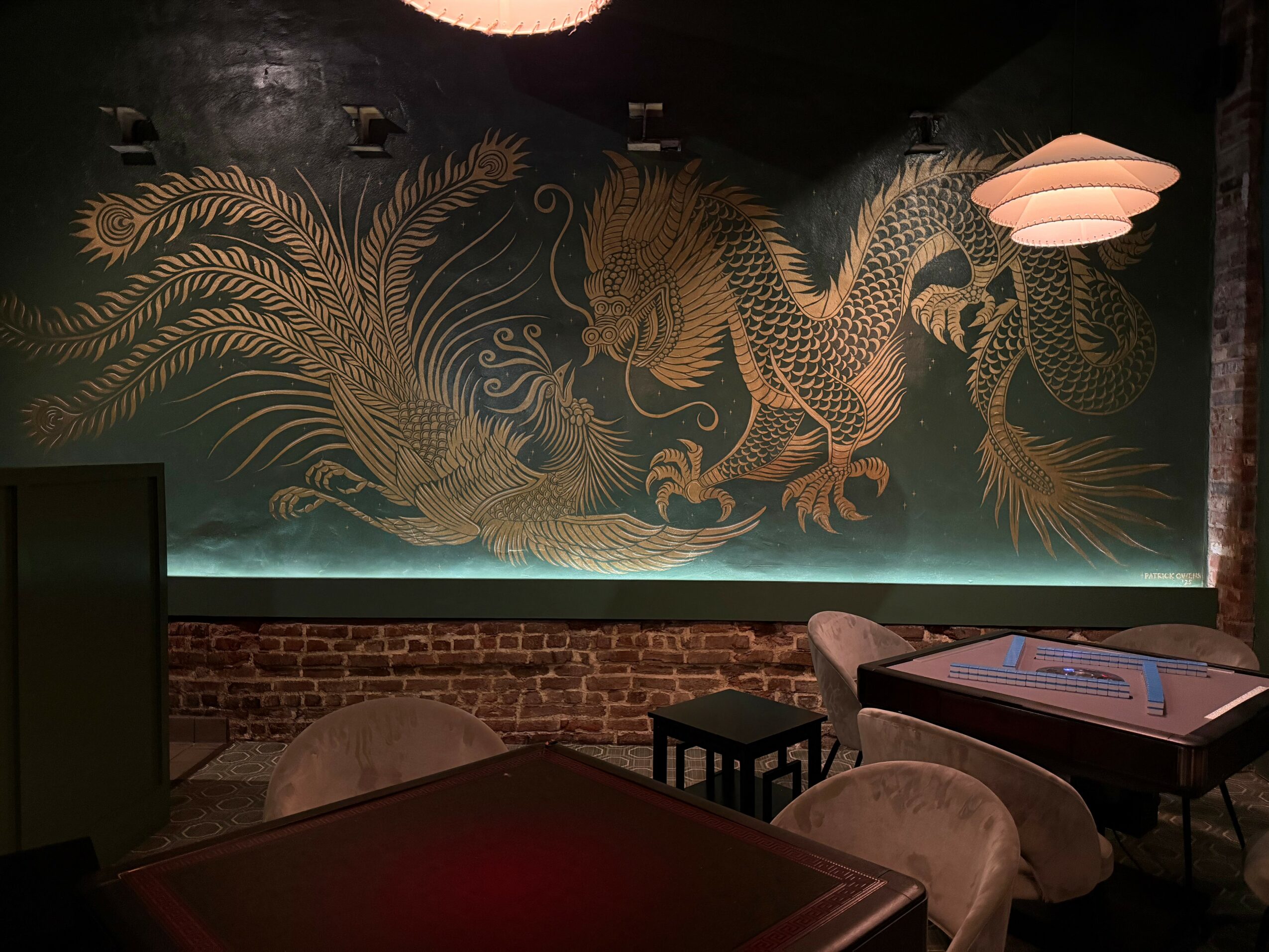 Decorative wall with a golden dragon and foliage design, illuminated by soft lighting, in a dimly lit room with tables and chairs.