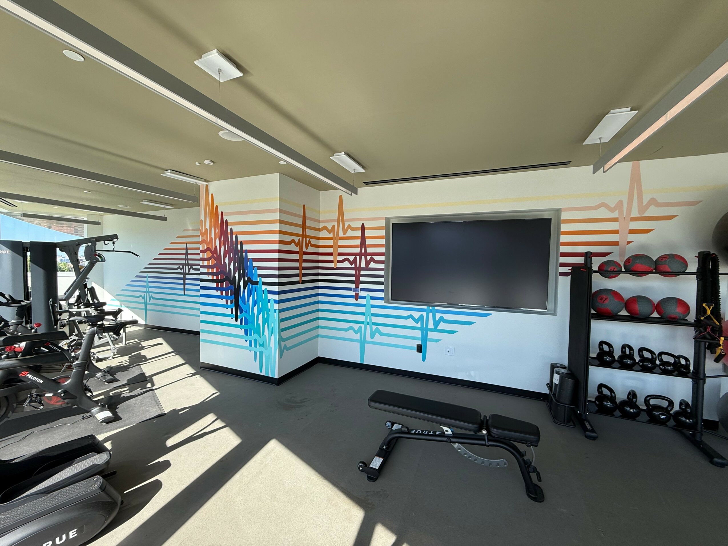 Gym interior with exercise equipment, a wall with colorful heartbeat line mural, and a large screen on the wall.