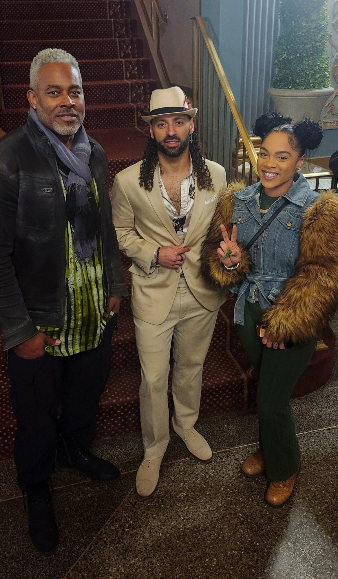 Three people standing indoors, smiling, with a staircase and window in the background.