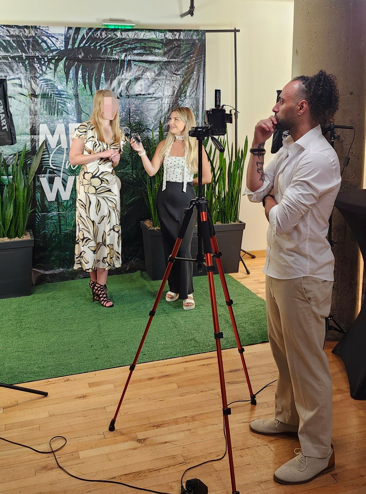 Three people in a studio with a green backdrop, two women talking and a man observing, camera on tripod, wooden floor.
