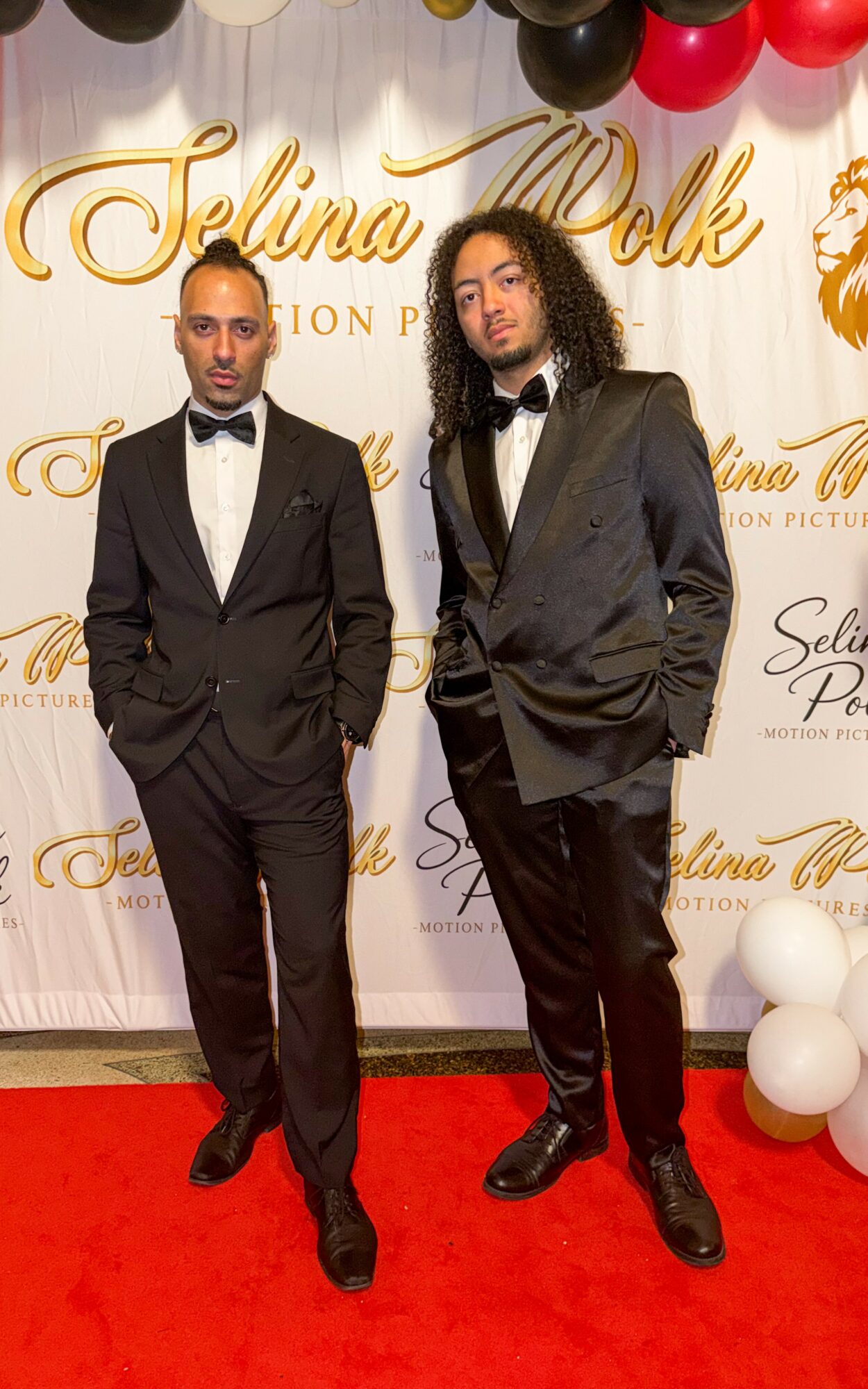 Two men in tuxedos standing on a red carpet in front of a backdrop with gold text and balloons.