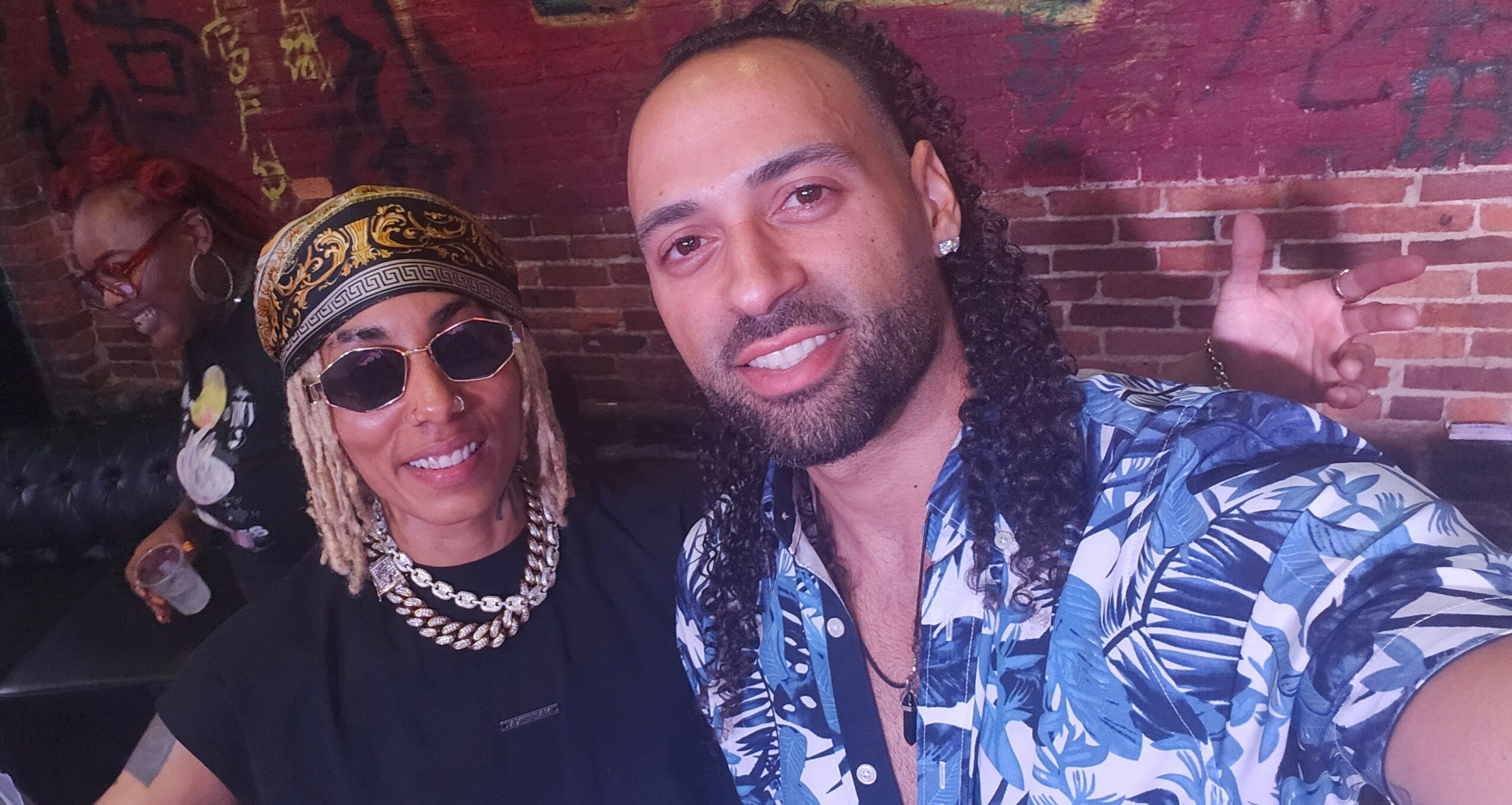Two people smiling, one wearing sunglasses and a patterned headscarf, the other with long curly hair and a patterned shirt, in front of a brick wall.