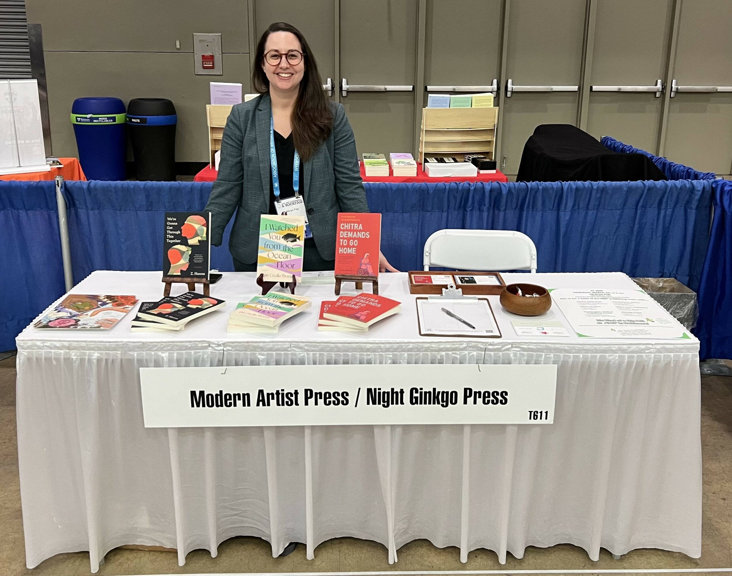 Woman standing behind a table with books and promotional materials at a booth, with a blue tablecloth and a sign reading 'Modern Artist Press / Night Ginkgo Press'.