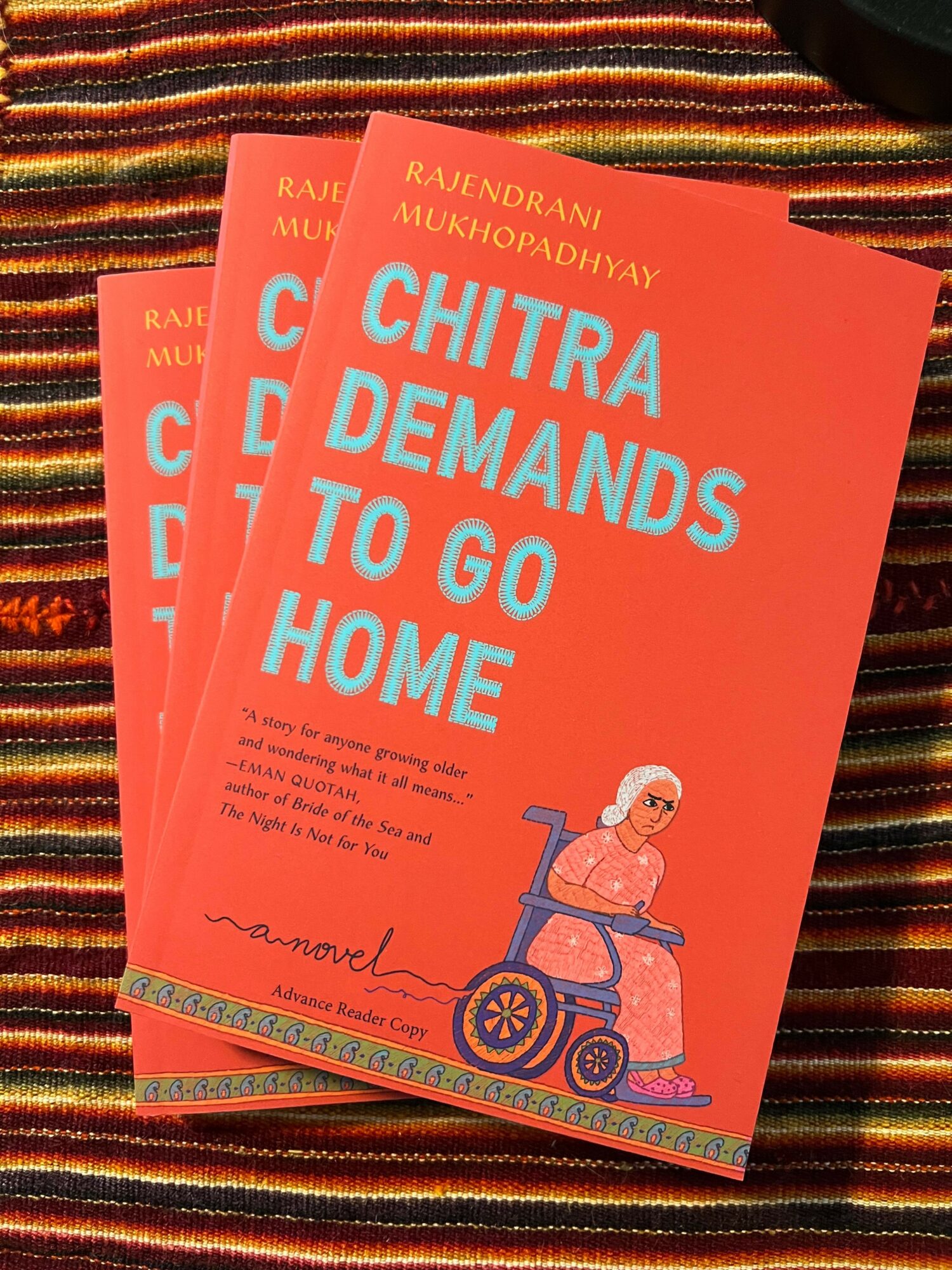 Two pink books titled 'Chitra Demands to Go Home' on a striped fabric surface.