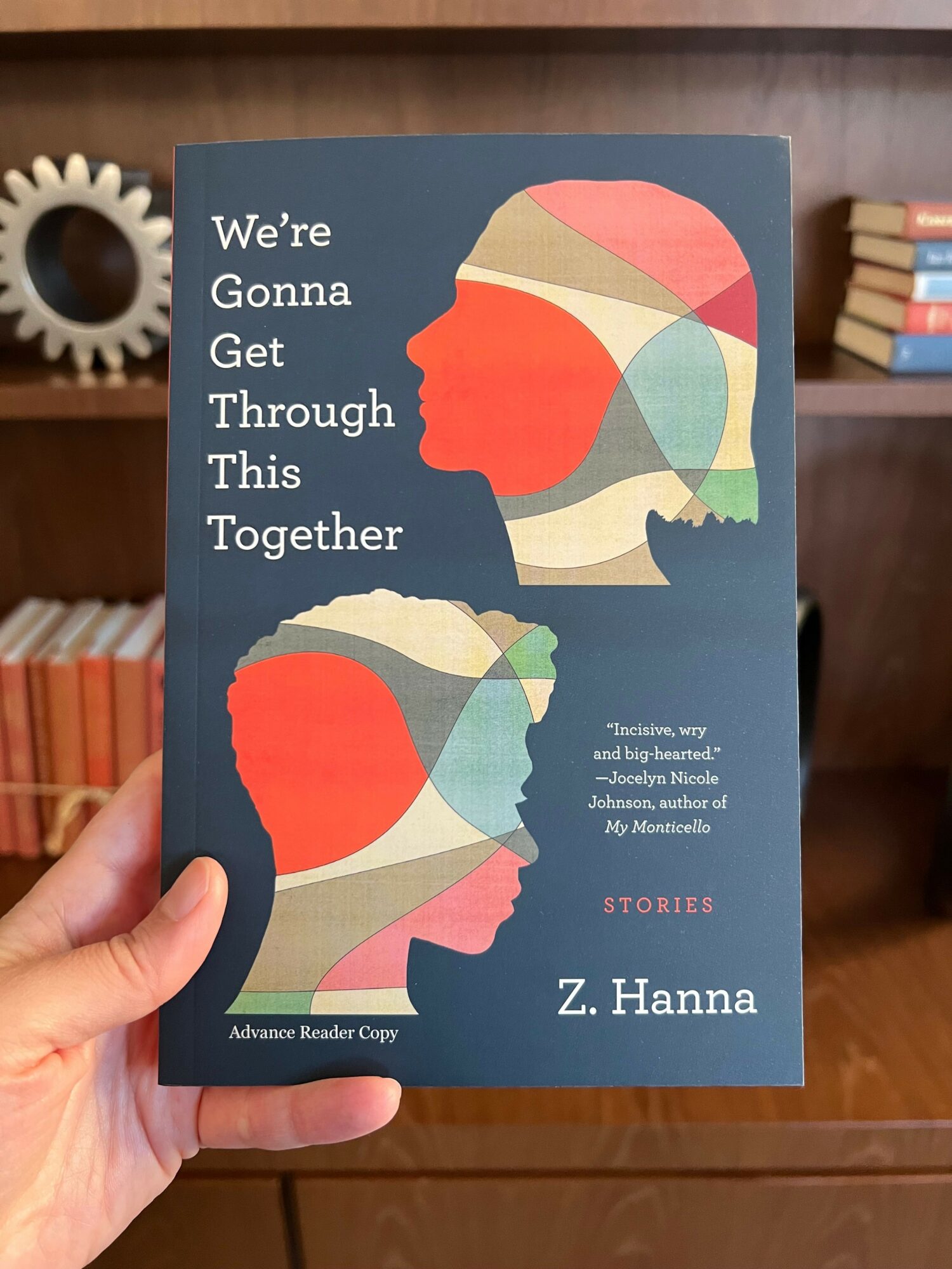 Book cover with colorful abstract profile silhouettes of a person and a child, title and author's name, background of bookshelves.
