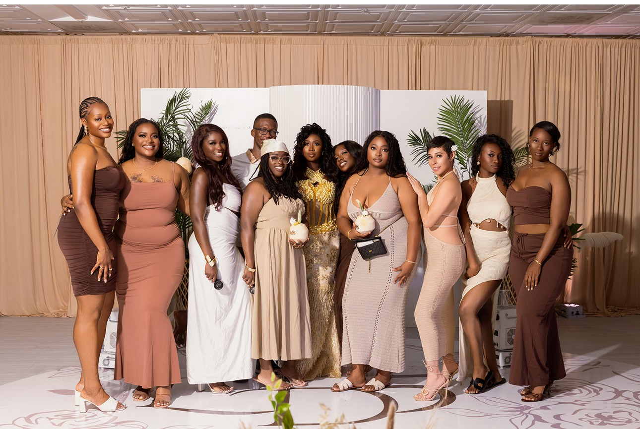 Group of ten women standing together indoors, some holding flowers, in front of a decorated backdrop with plants.