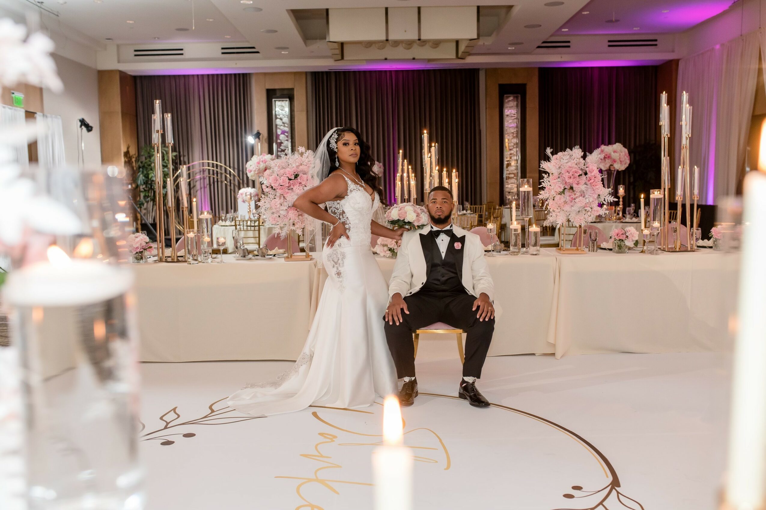 A bride in a white wedding dress and a groom in a tuxedo pose in a decorated reception hall with pink flowers and candles.