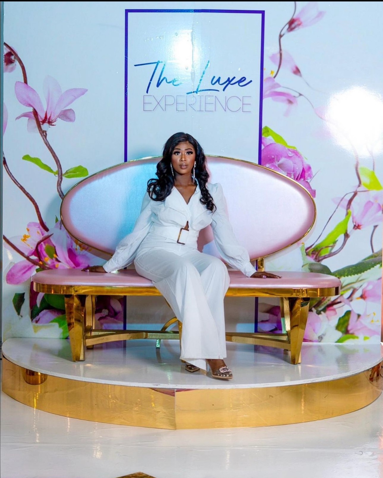 Woman in white outfit sitting on a pink and gold bench with floral background and a sign that reads 'The Luxe Experience'.
