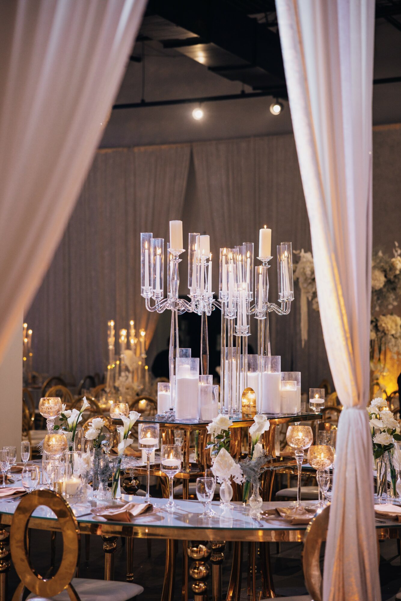 Elegant banquet table with tall candleholders, floral arrangements, and tableware, seen through curtains in a decorated event space.