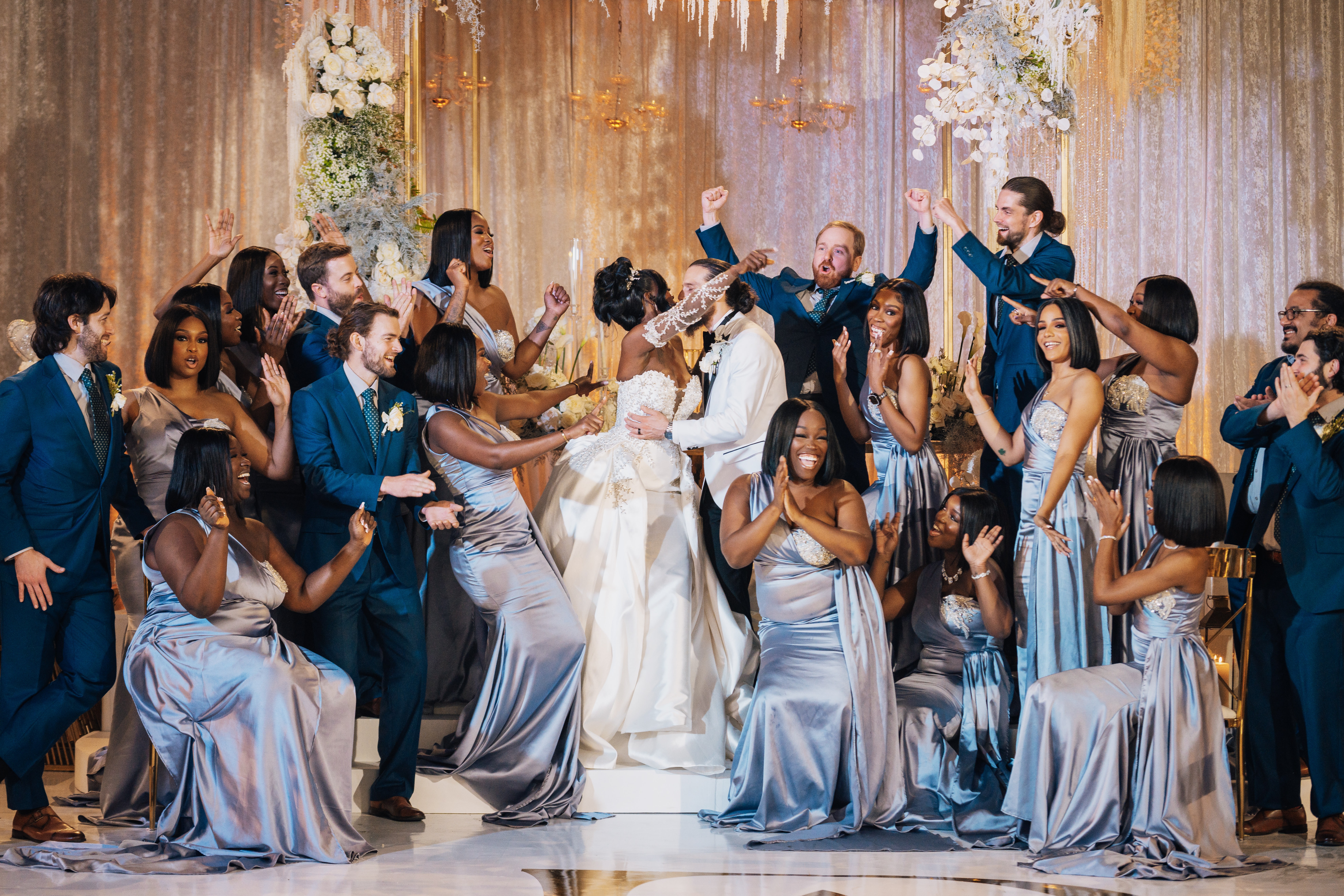 Group of people dancing and celebrating at a wedding reception with floral decorations and elegant attire.