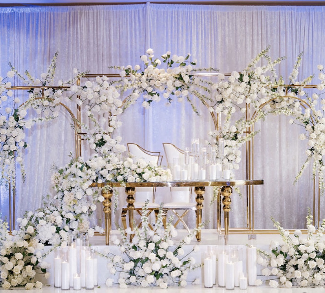 Elegant wedding stage with white flowers, gold accents, and two chairs, decorated with floral arrangements and candles.