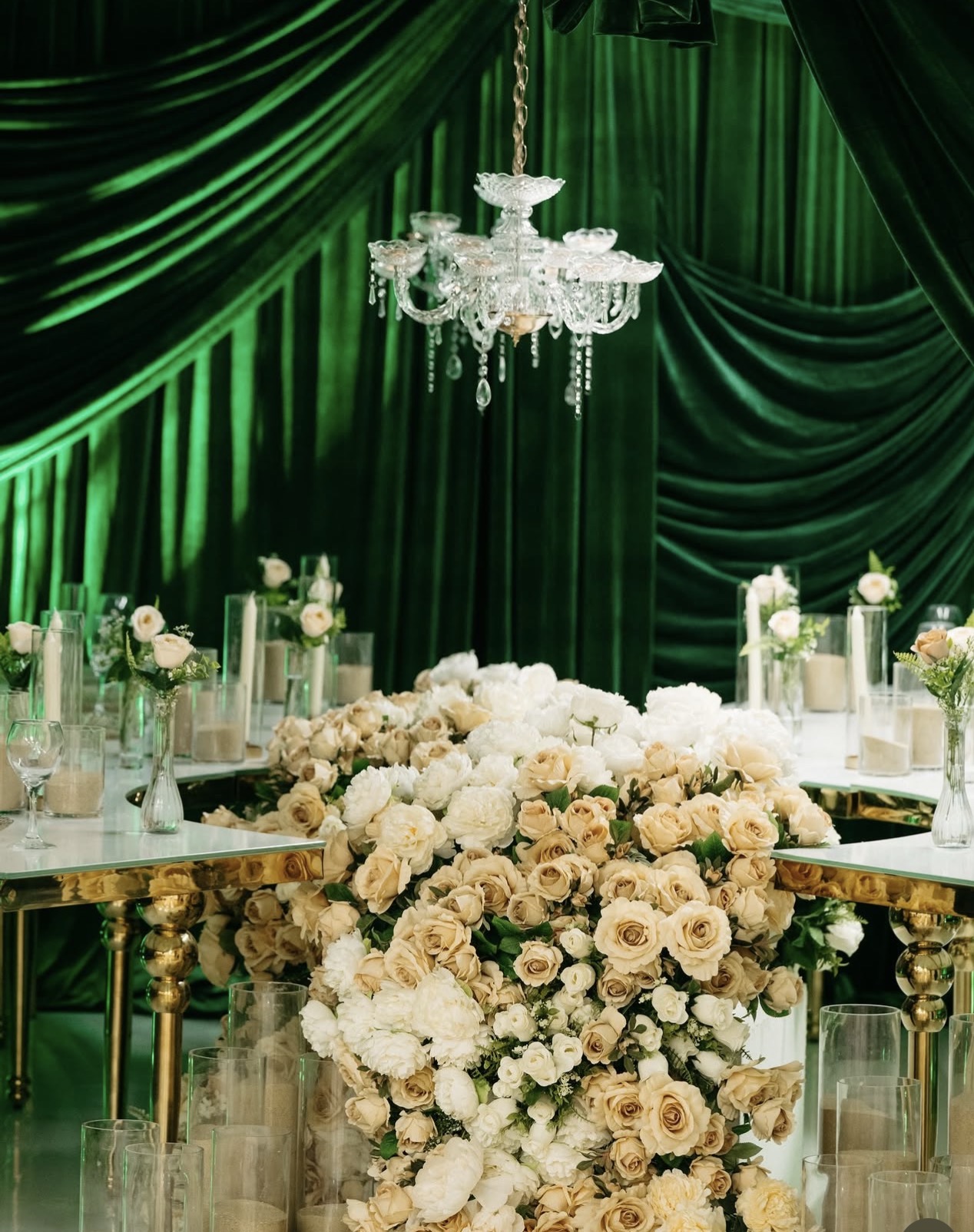 Elegant event table with floral centerpiece, green draped backdrop, and chandelier, decorated with candles and glassware.