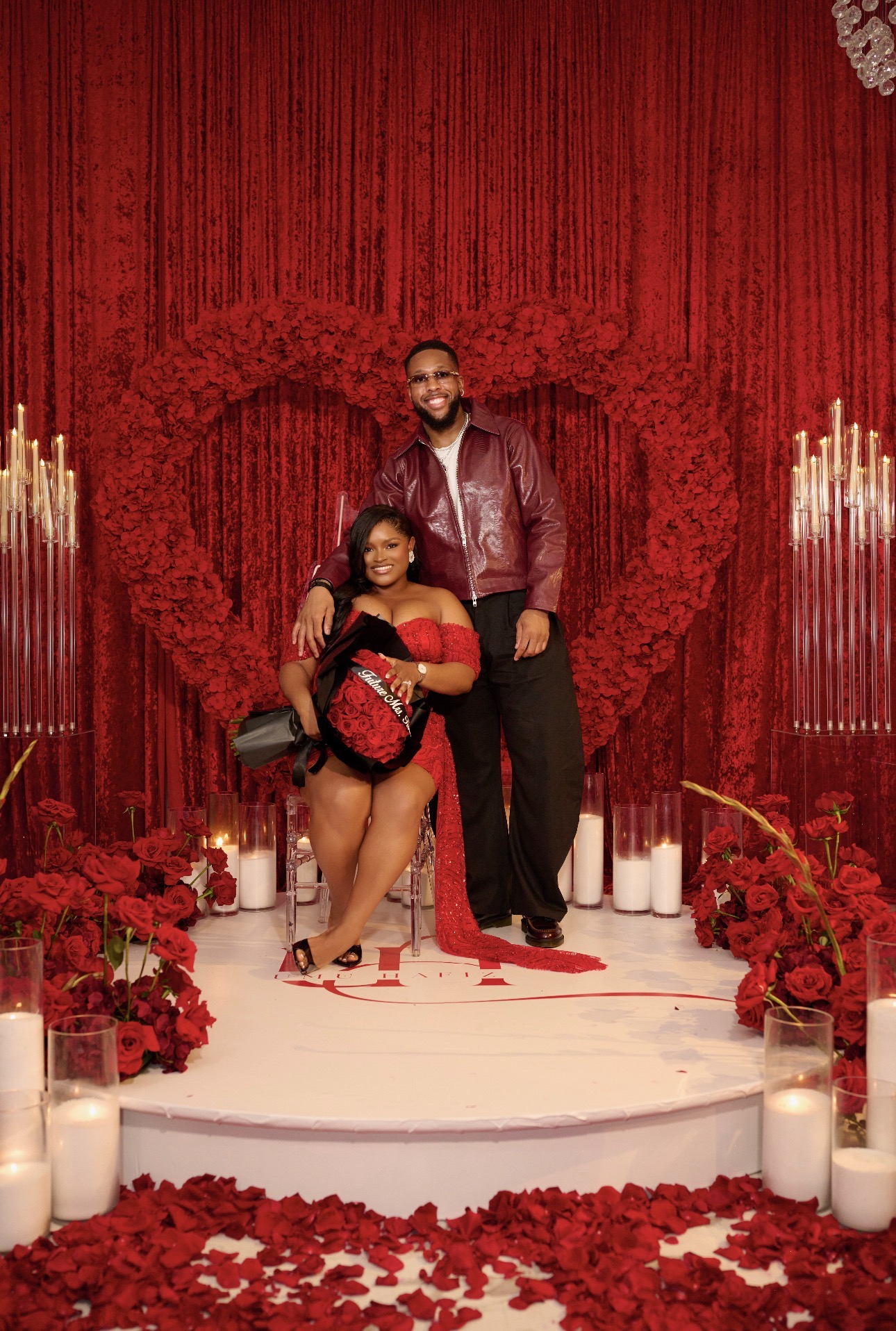 A couple stands on a decorated stage with a heart-shaped backdrop, surrounded by candles and red flowers, celebrating love.