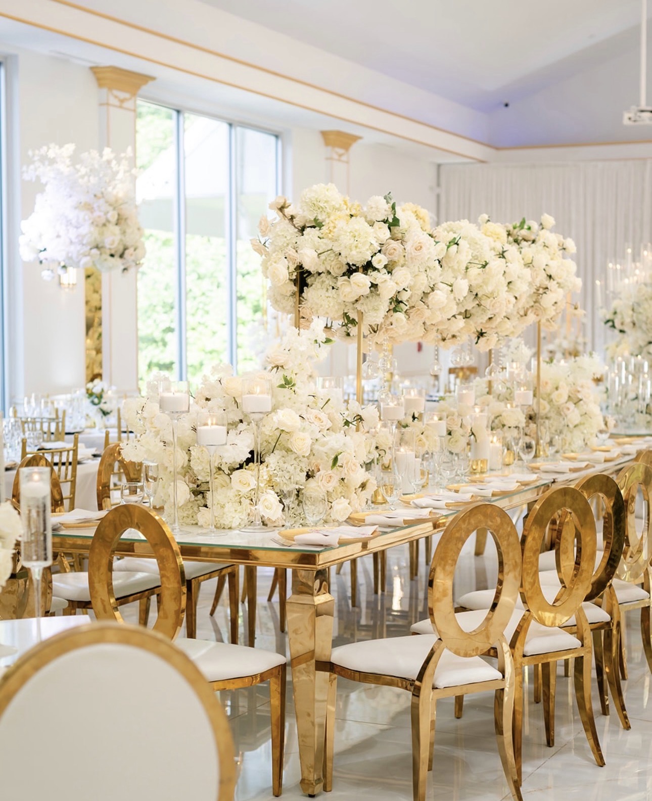 Elegant dining table with white floral centerpieces and gold chairs in a bright, decorated room with large windows.