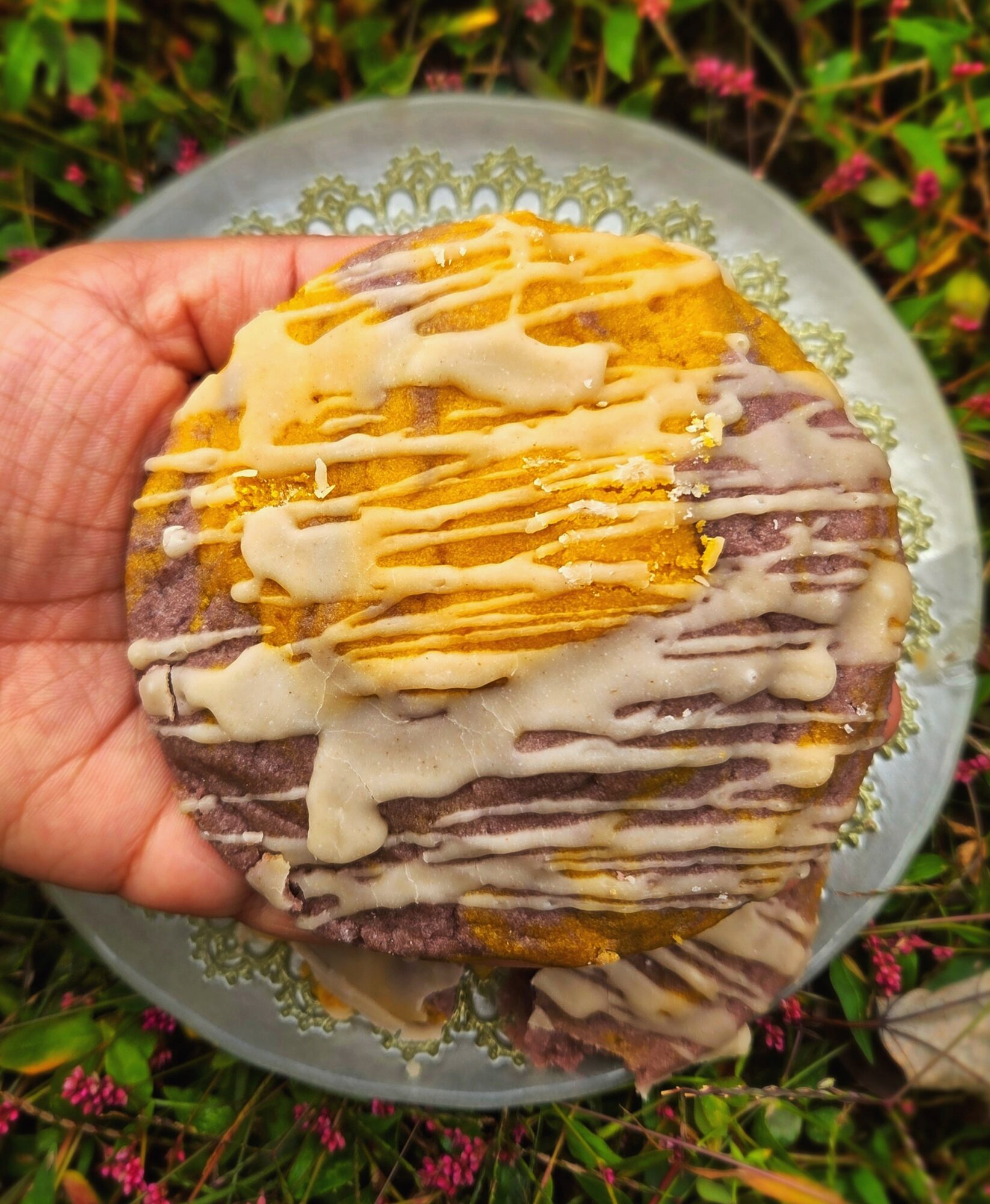 Hand holding a plate with layered snack topped with yellow and white sauces, outdoors with greenery and flowers.