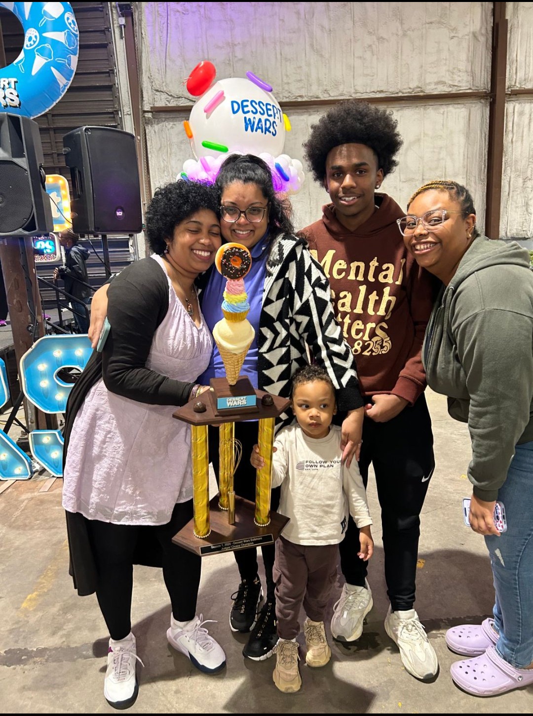 Four people and a child stand together indoors, smiling, with a colorful balloon and decorations in the background.