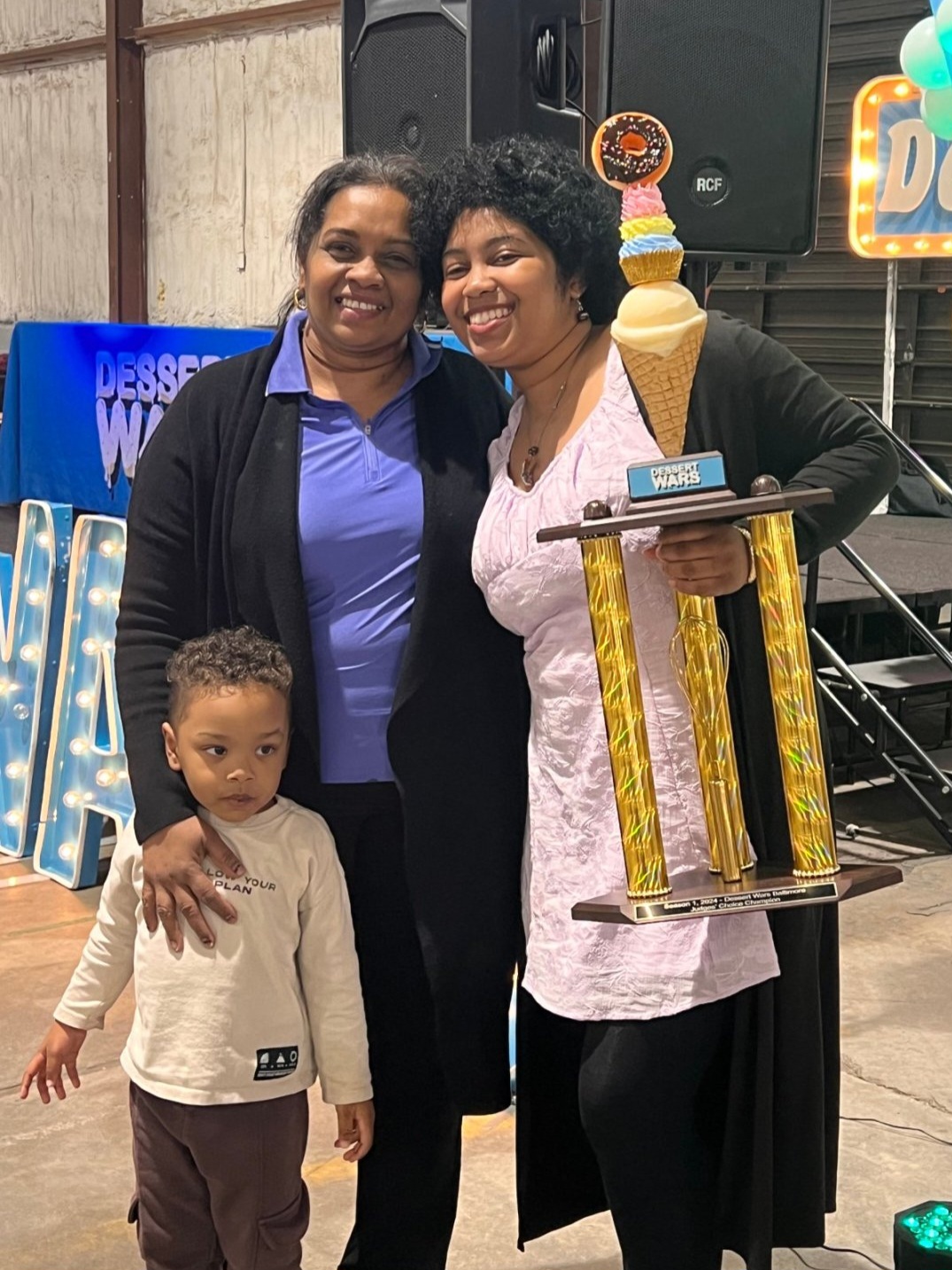 Three people, two women and a child, standing together indoors, one woman holding a large trophy with a tall ice cream cone on top.